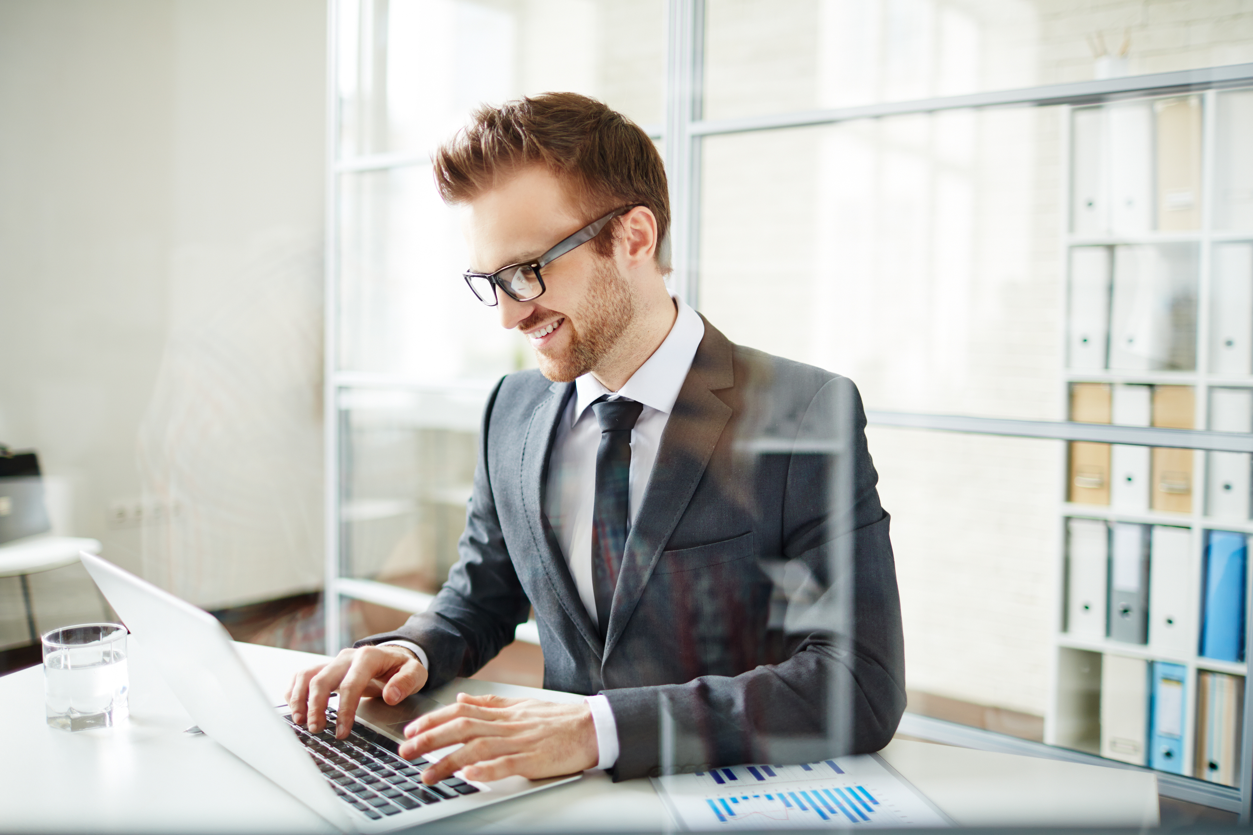 Ein Mann mit rot-braunen Haaren und Brille sitzt im Anzug am Schreibtisch und schaut auf einen geöffneten Laptop.