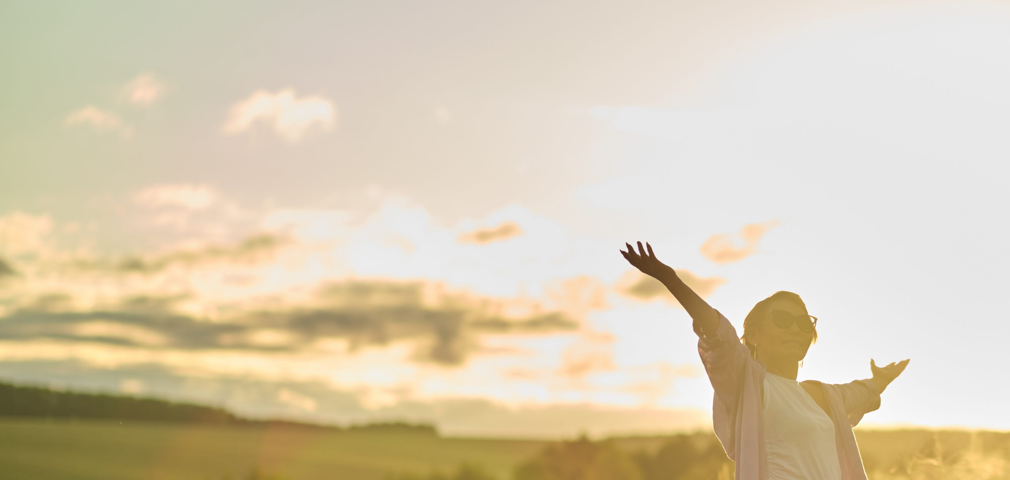 Eine Frau steht in einem Feld und hebt ihre Arme in die Luft. Die Sonne steht tief am Himmel und taucht alles in ein goldenes Licht.