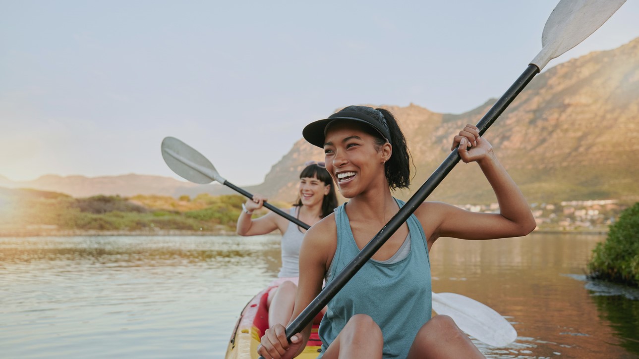 Zwei Frauen sitzen im Kajak auf einem See und halten lachend die Paddel in den Händen. Im Hintergrund ist eine Berglandschaft und eine Stadt am Ufer.