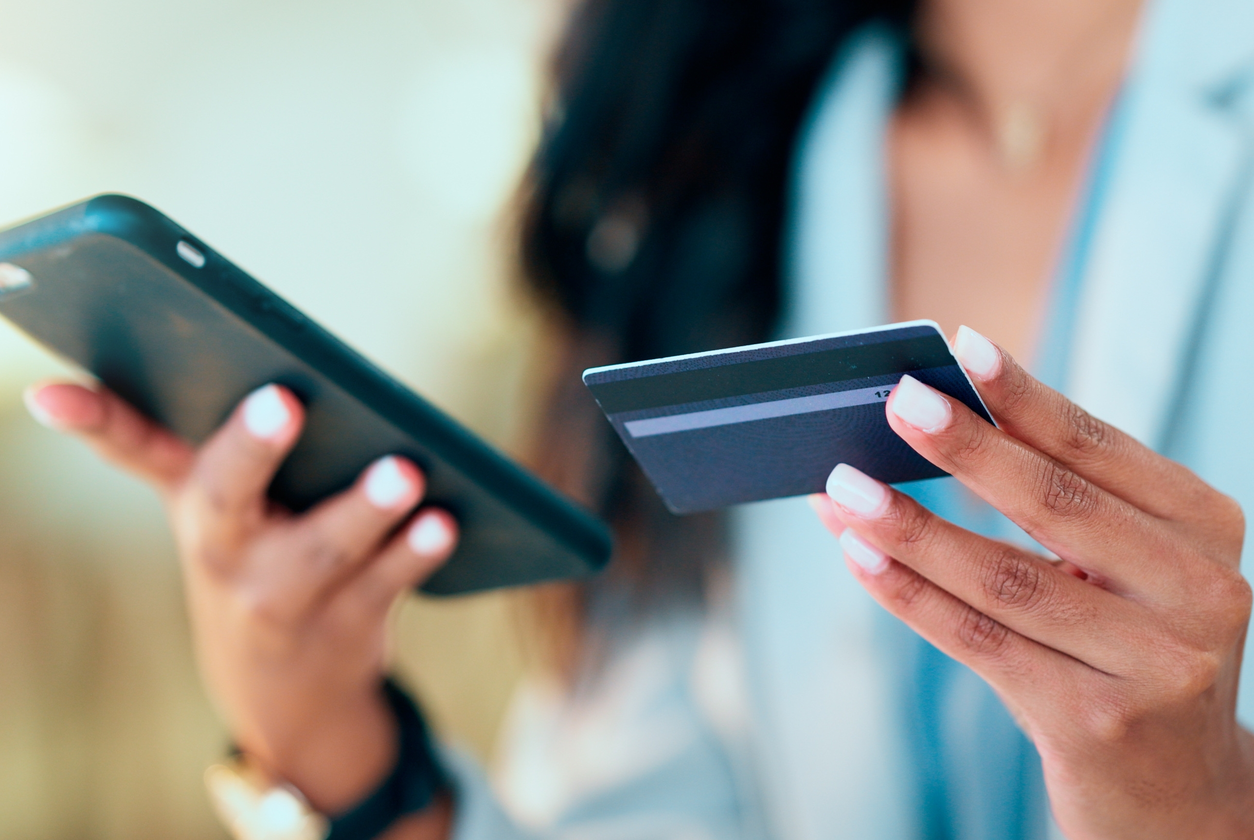 Eine Frau hält ein Smartphone und eine Kreditkarte in der hand. Sie trägt einen hellblauen Blazer und ein hellblaues Hemd und hat lange schwarze Haare.