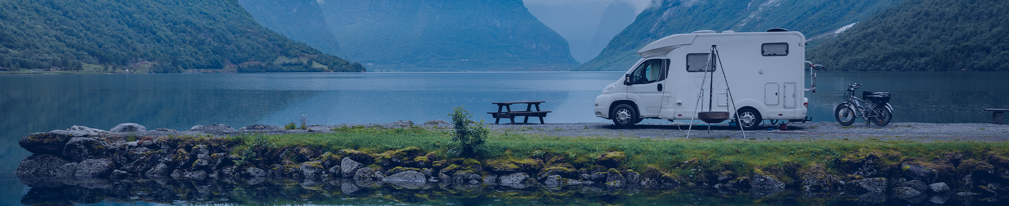 Das Bild ist mit einem blauen Filter hinterlegt. In der Mitte sieht man einen Mann im Anzug am Schreibtisch sitzen. Vor ihm liegen diverse Papiere und im rechten Bildrand steht ein Laptop.