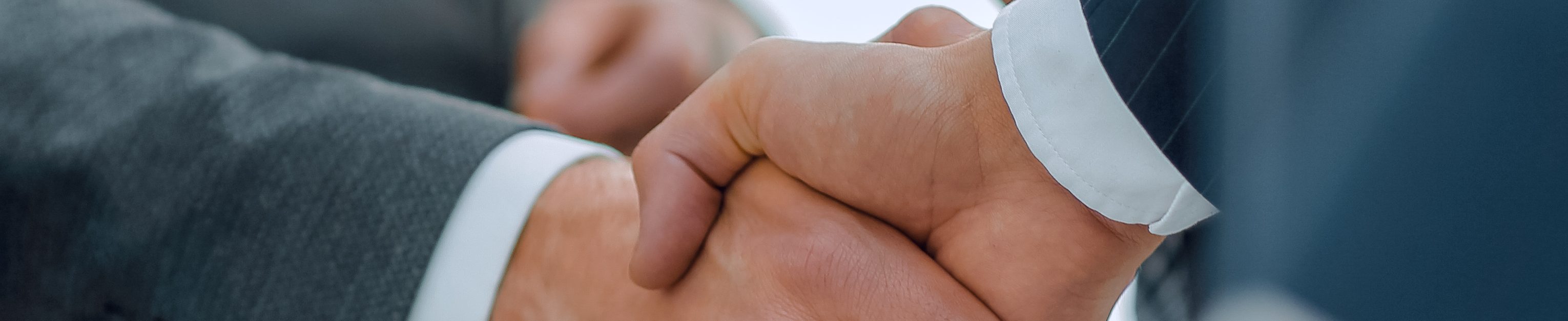 Zwei Männer machen in der Mitte des Bilds einen Handschlag. Der linke trägt einen grauen Anzug, der rechte einen blauen Anzug, beide darunter ein weißes Hemd. Im Bild sind nur die Hände ersichtlich.