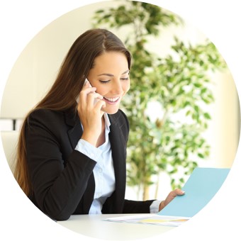 Eine Frau in Hemd und Blazer sitzt am Tisch und telefoniert während sie auf eine geöffnete Arbeitsmappe schaut und lächelt.