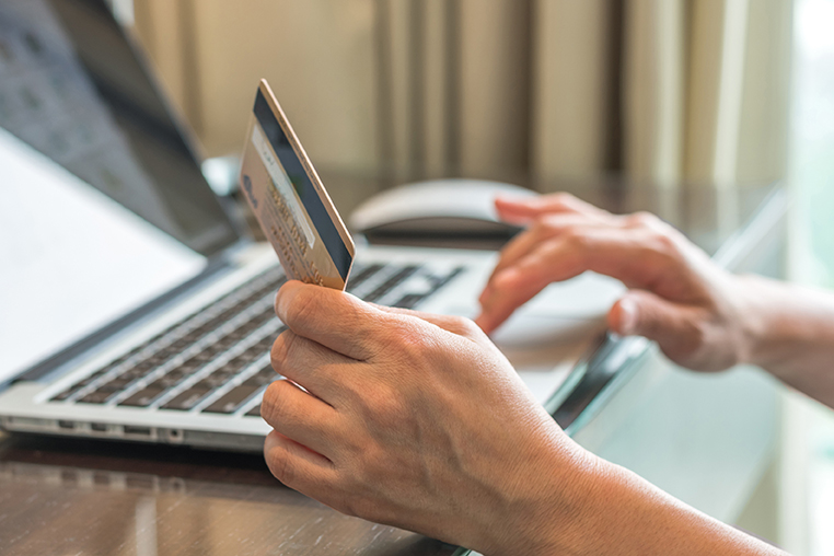 Eine Person sitzt an einem Tisch und tippt mit der einen Hand auf einem Laptop und hält mit der anderen Hand eine Kreditkarte.