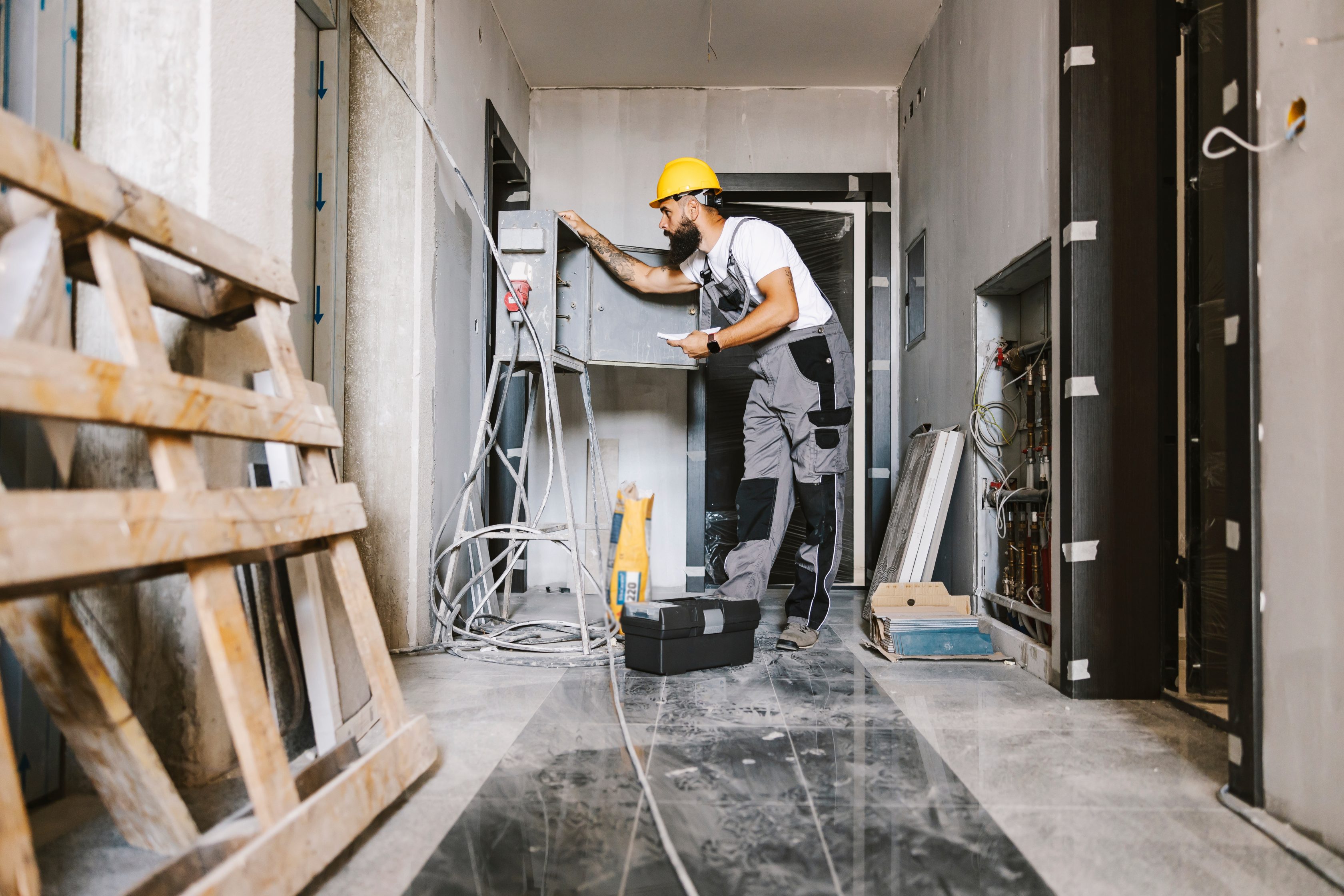 Ein Handwerker mit gelben Helm steht in einem renoviertem Raum und schaut in einen geöffneten Sicherungskasten.