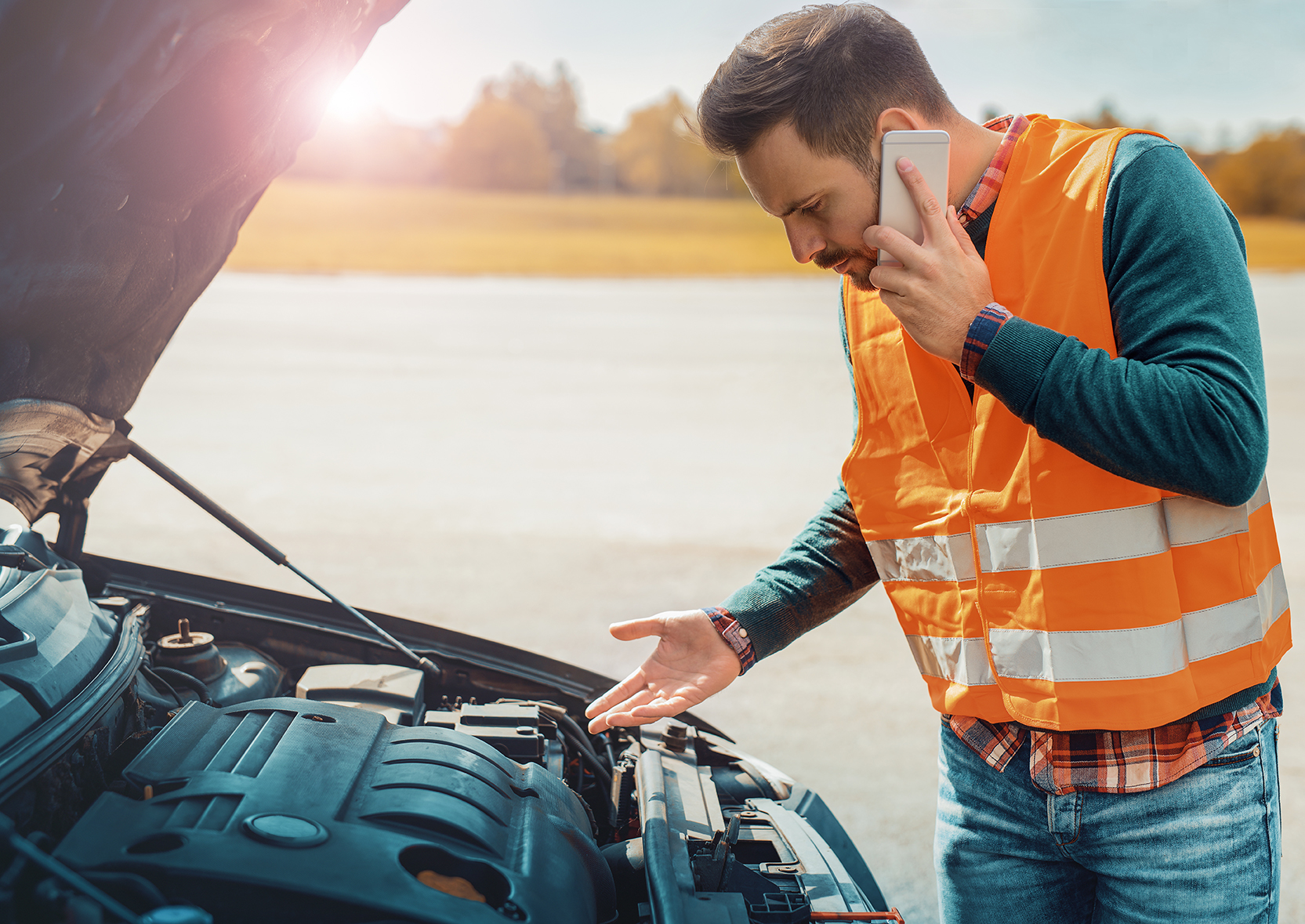 Ein Mann mit Handy in der linken Hand trägt eine orangene Warnweste und steht fragend bzw. mit der rechten Hand auf eine geöffnete Motorhaube zeigend. Im Hintergrund ist eine Straße und dahinter Landschaft zu sehen.