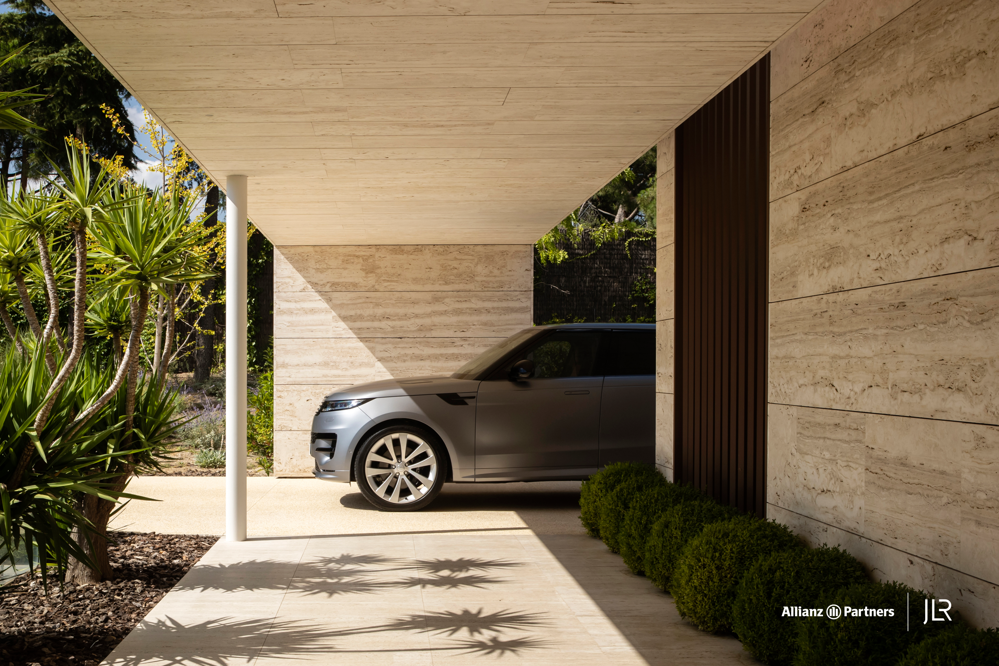Ein grauer SUV steht vor einem modernen Haus mit beiger Fassade. Links und rechts sind grüne Pflanzen eingepflanzt.