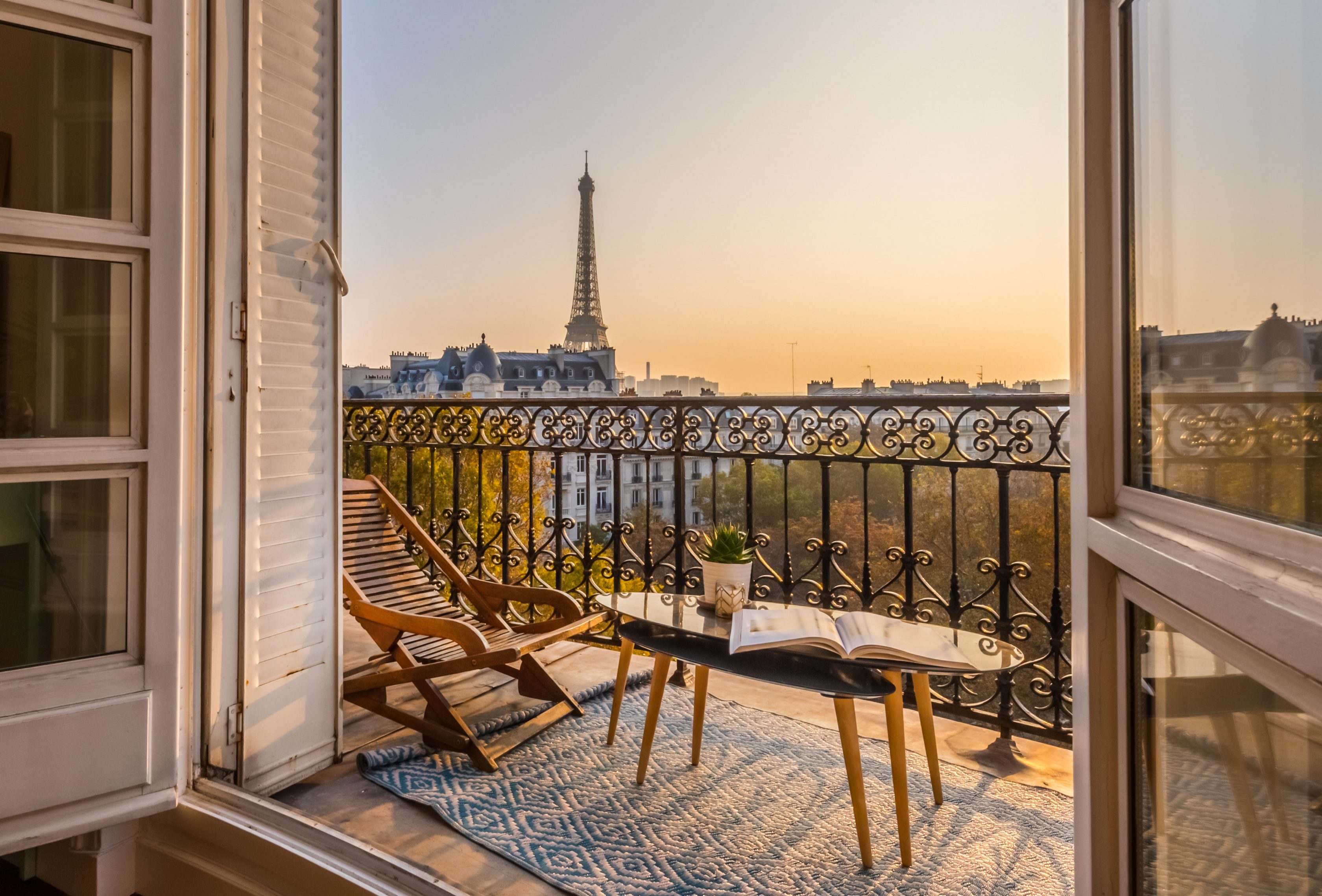 Eine Balkonansicht in Paris mit Blick auf den Eiffelturm bei Sonnenuntergang. Auf dem Balkon stehen ein Holzliegestuhl, ein Glastisch mit einer Pflanze, einer Kerze und einem Buch.