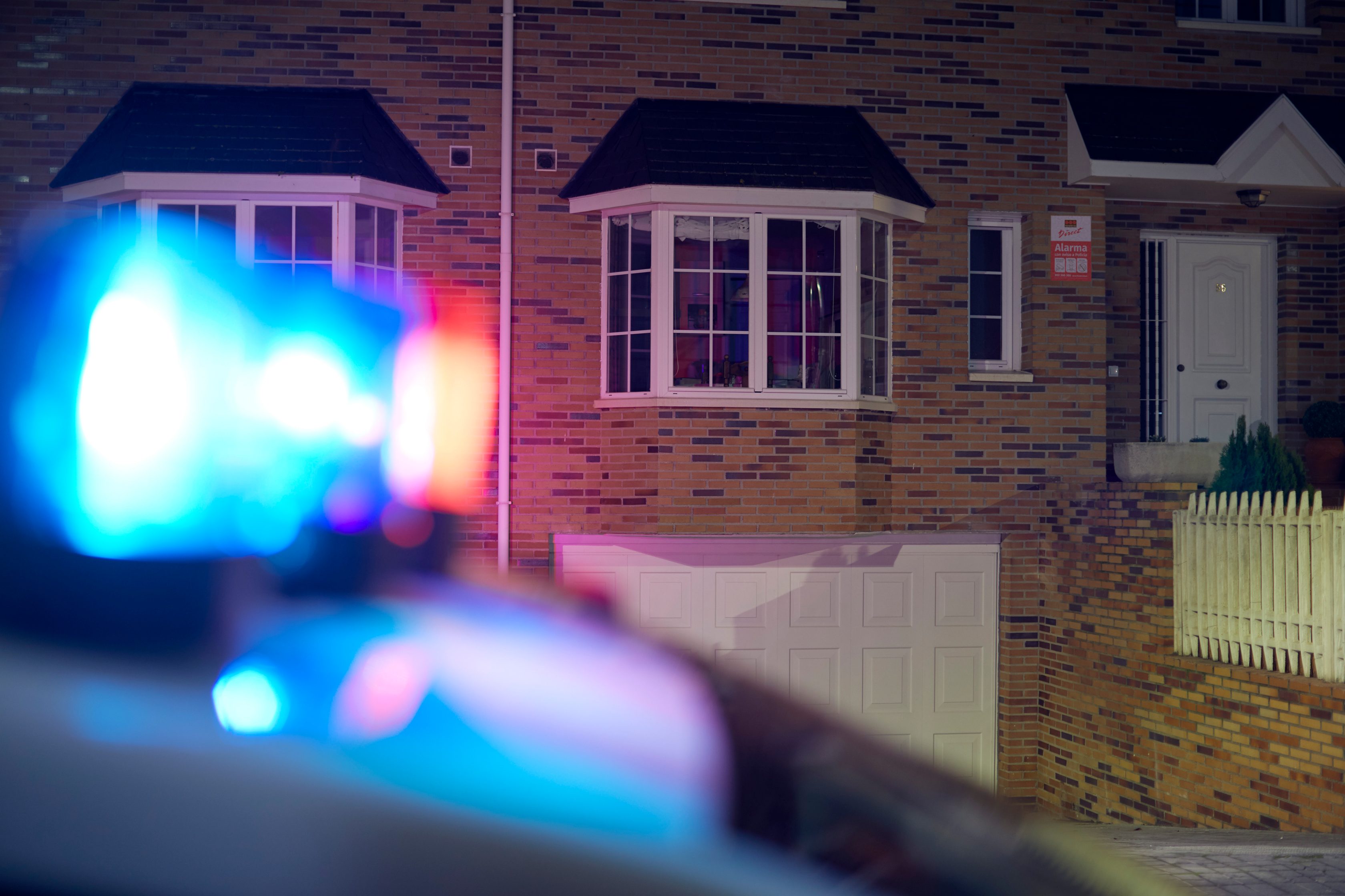 Frontansicht eines Backsteinhauses mit heller Haustür, zwei hellen Fenstern und blinkenden Lichtern eines Polizeiautos im Vordergrund.