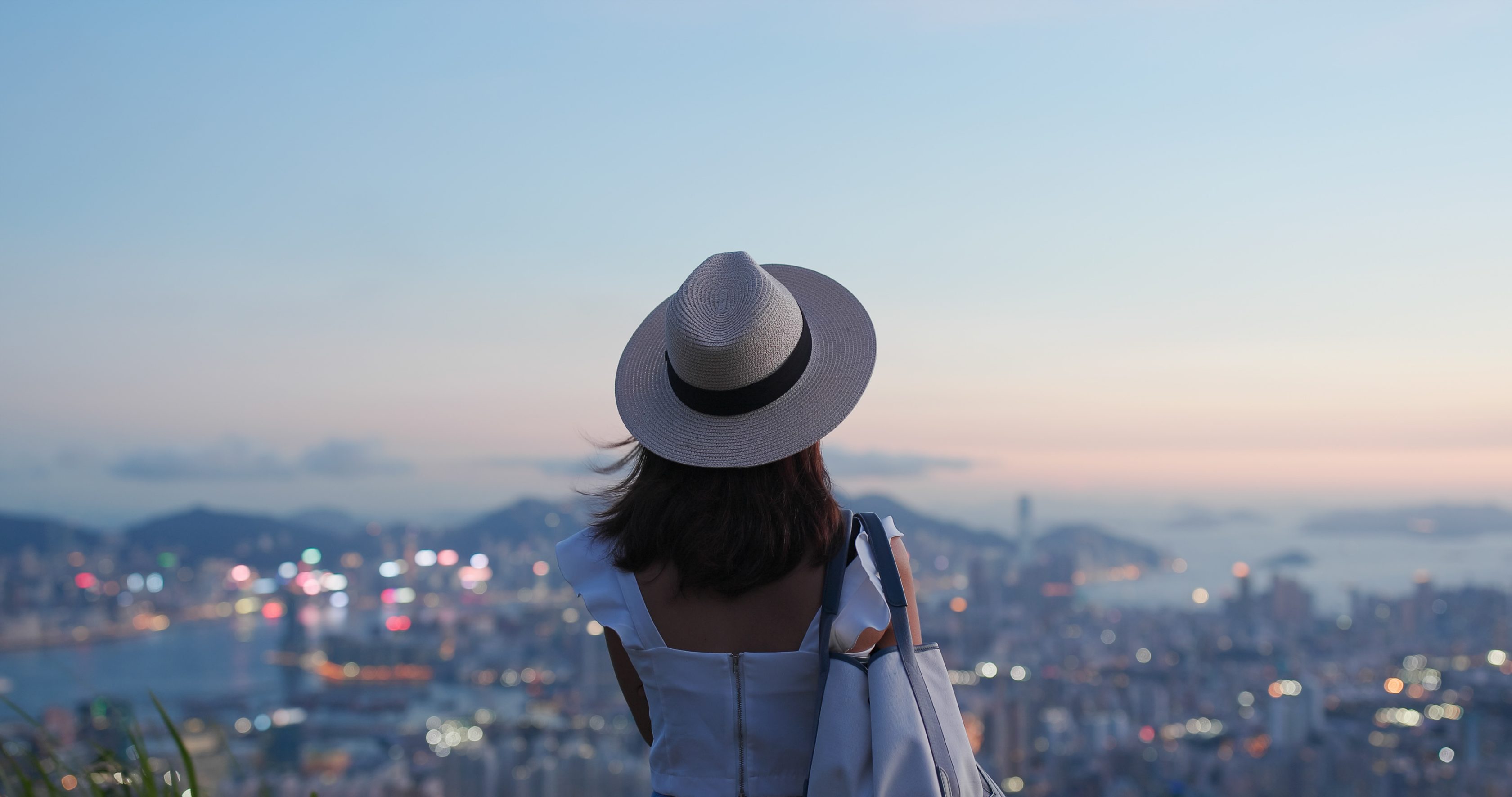 Eine Frau in weißem Outfit mit breitkrempigem Hut blickt auf eine Stadtlandschaft bei Dämmerung, im Hintergrund Gebäude und Wasser.