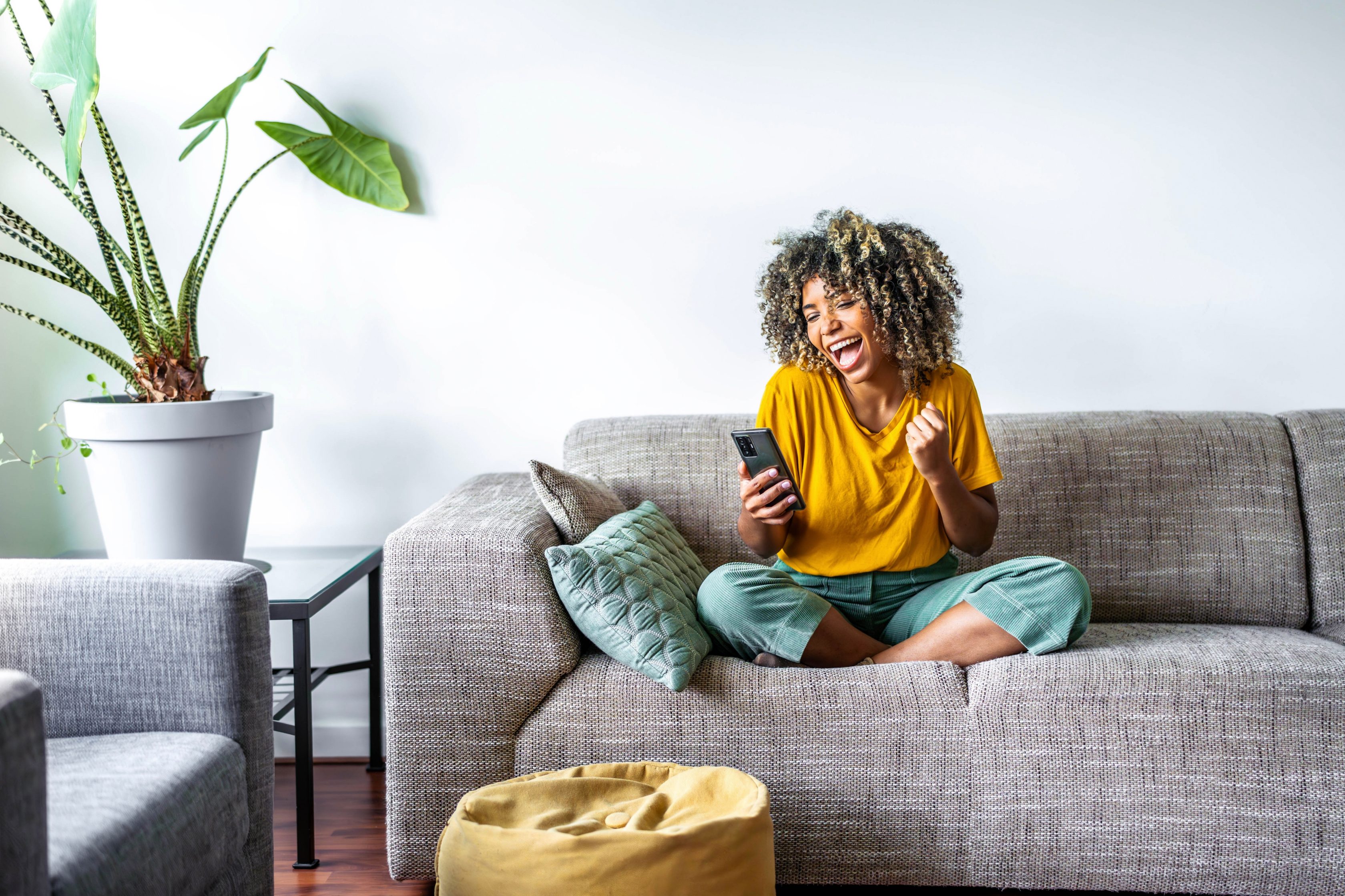 Eine Frau sitzt mit dem Smartphone in der Hand auf einem Sofa in einem Wohnzimmer und schaut lachend auf das Handy.