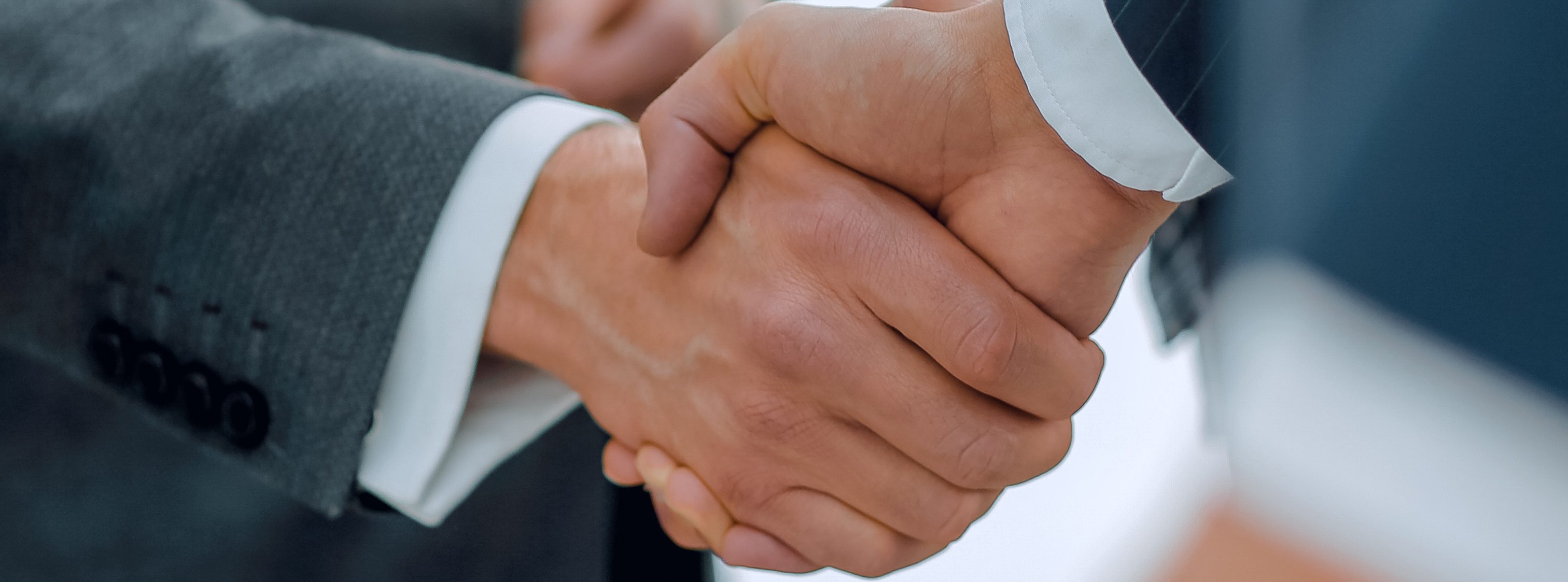 Zwei Männer machen in der Mitte des Bilds einen Handschlag. Der linke trägt einen grauen Anzug, der rechte einen blauen Anzug, beide darunter ein weißes Hemd. Im Bild sind nur die Hände ersichtlich.