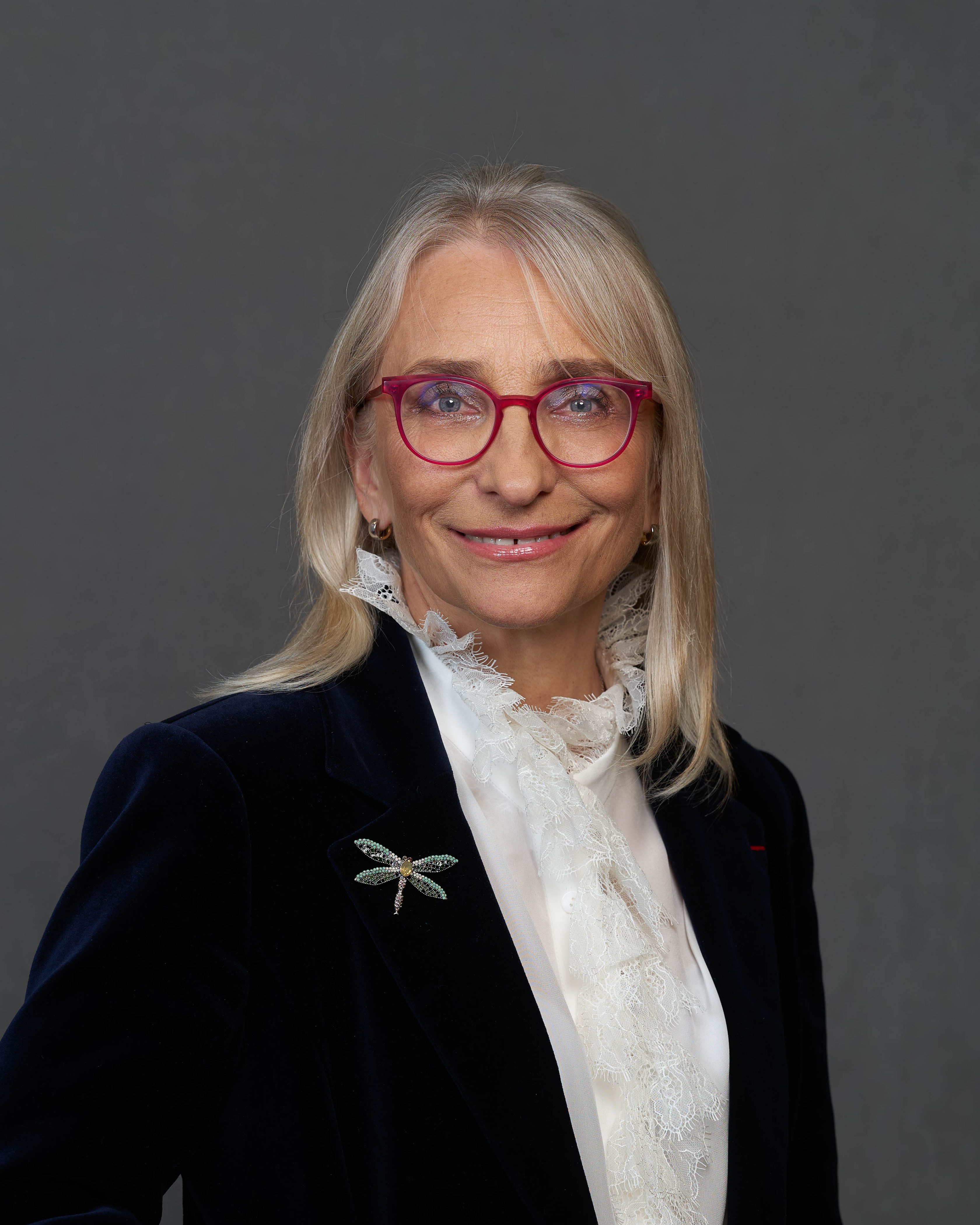 Porträt von Ida Luka-Lognoné. Eine Frau mit roter Brille trägt eine weiße Schluppenbluse mit Rüschen, darüber einen schwarzen Blazer. An diesem ist auf der linken Seite eine Glitzerlibelle als Anstecknadel befestigt. Ida lächelt in die Kamera und zeigt dabei leicht ihre Zähne.