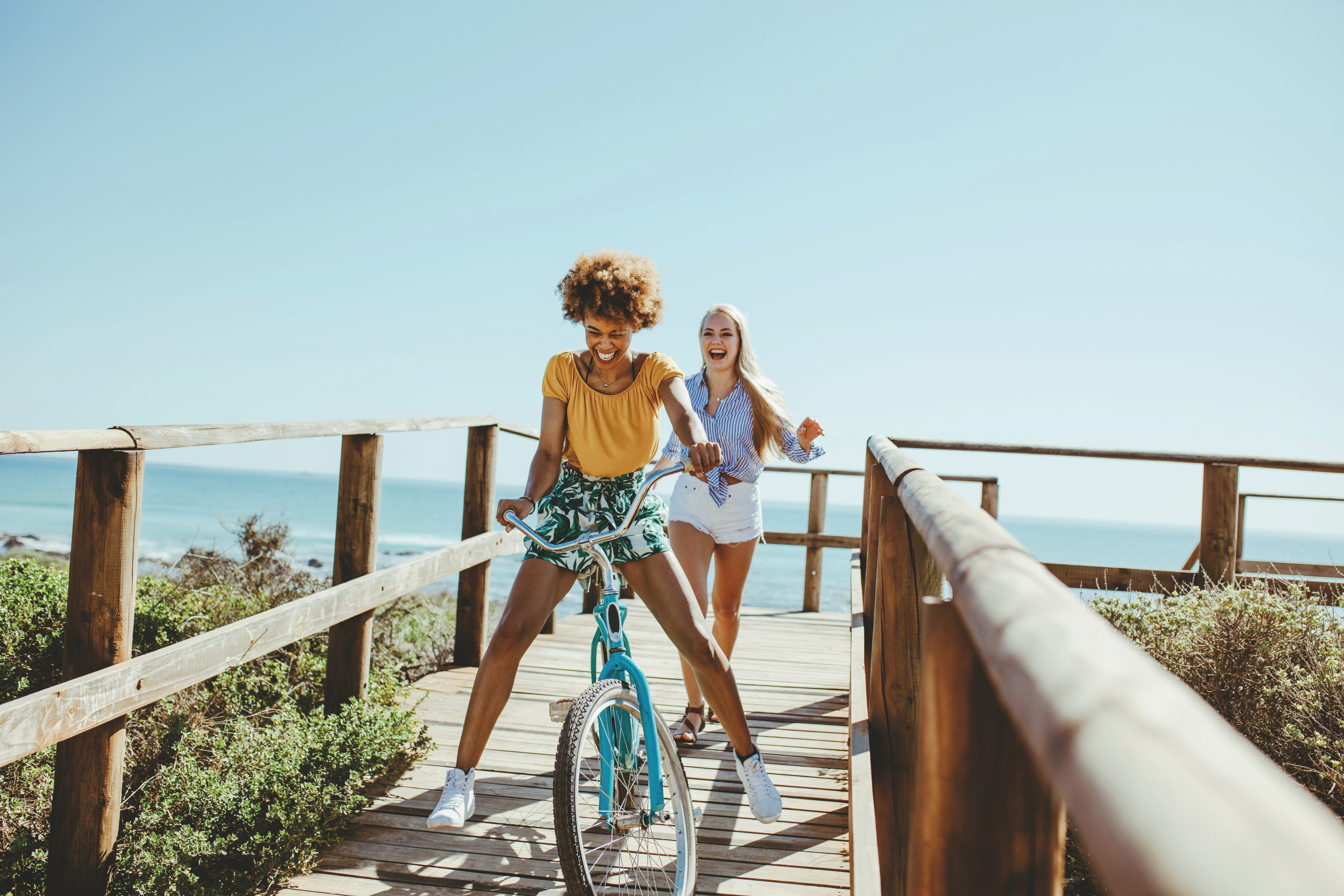 Allianz Partners Deutschland – Neue Reiseschutztarife: Zwei fröhlich lachende Personen, eine auf einem Fahrrad und eine zu Fuß, bewegen sich entlang eines Holzstegs mit Blick auf das weite Meer unter klarem Himmel.