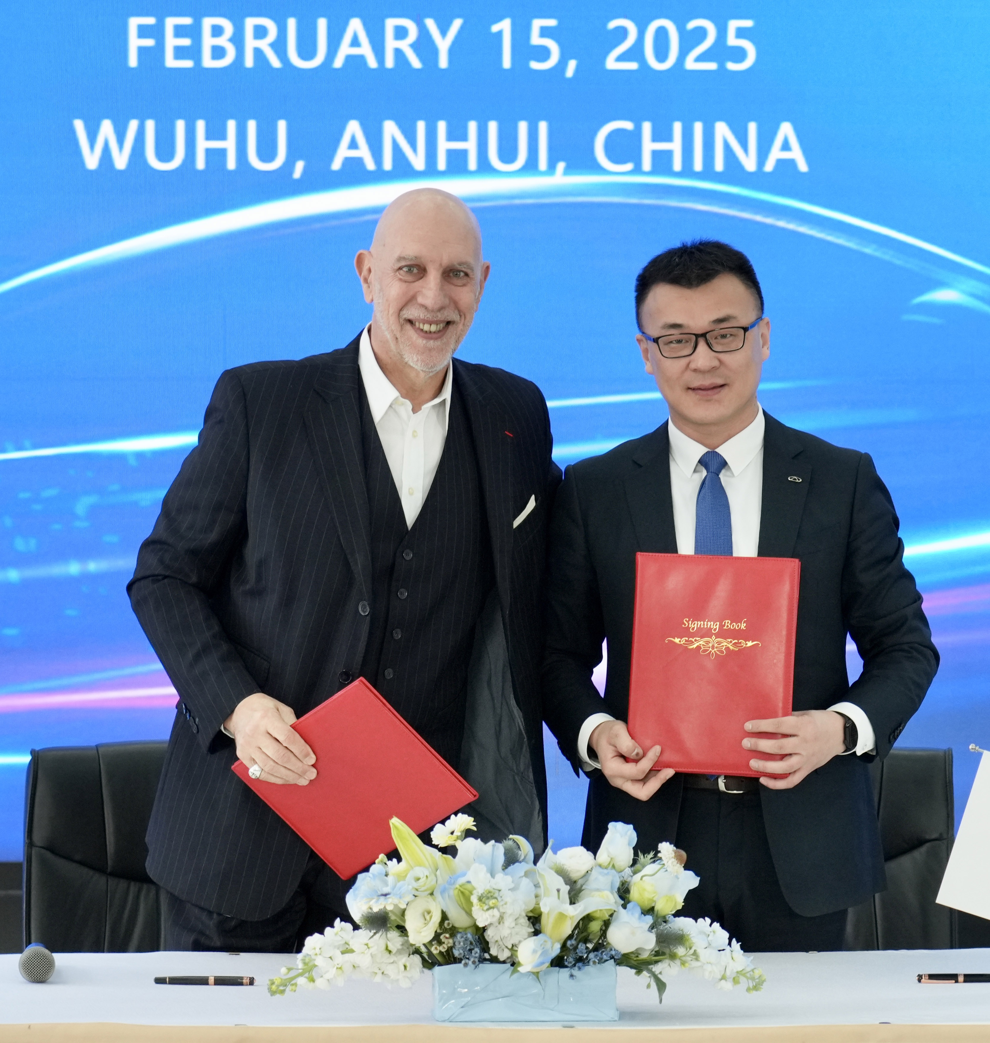 Ein Foto von Jean-Marc Pailhol und Charlie Zhang. Beide Männer stehen im schwarzen Anzug mittig im Bild unt halten die roten Ordner für die Vertragsunterlagen der Partnerschaft in der Hand. Vor ihnen sind weiße Blumen zu sehen, im Hintergrund eine blaue Wand und die Schrift "February 15, 2025. Wuhu, Anhui, China". 
