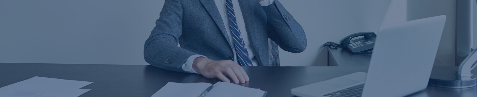 Eine Person in einem grauen Anzug sitzt an einem Schreibtisch mit Telefon, Laptop und Dokumenten.