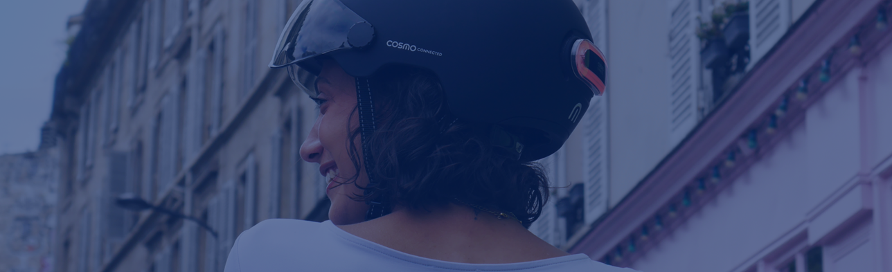 Eine Person trägt einen Fahrradhelm mit „Cosmo Connected“-Branding vor einem urbanen Hintergrund mit Straßen und Gebäuden.