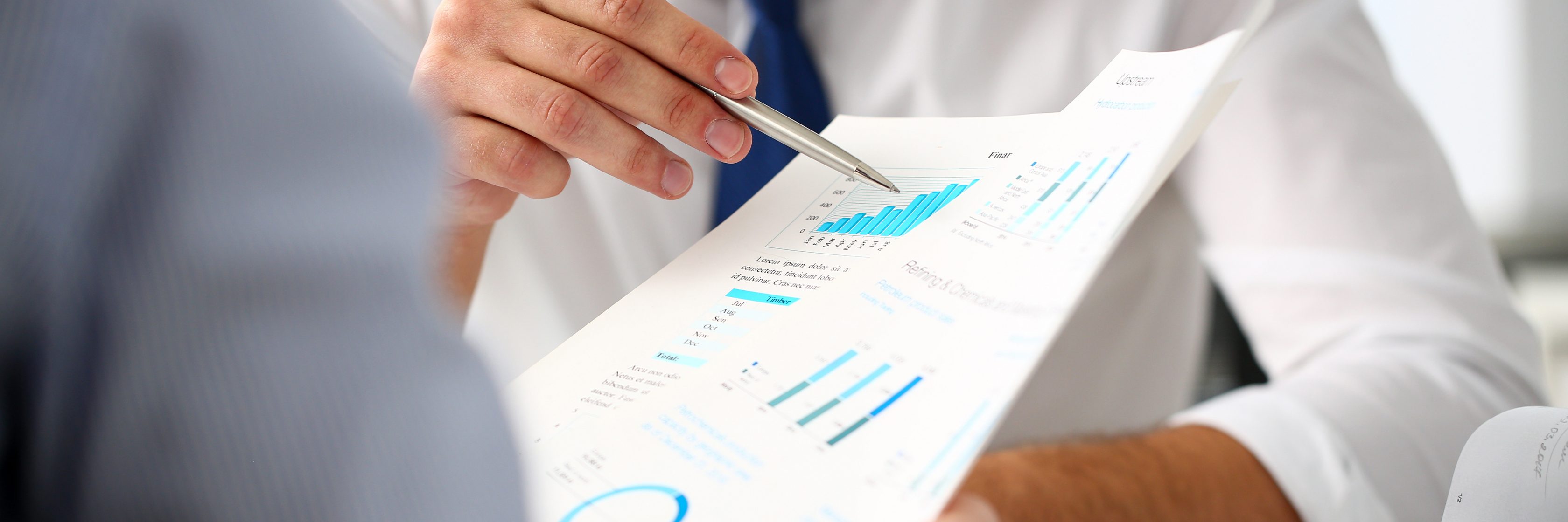 Das Bild zeigt zwei Männer in Hemden, links hellblau, rechts weiß, es sind aber nur links der rechte Arm des Mannes und rechts die linke Hand, der Oberkörper und der rechte Arm des Mannes zu sehen. Letzerer zeigt mit einem Stift in der linken Hand auf Dokumente mit diversen Grafiken.