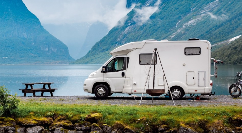 Das Bild zeigt ein weißes Wohnmobil vor einem Seepanorama mit Bergen im Hintergrund. Es ist bewölkt regnerisch.
