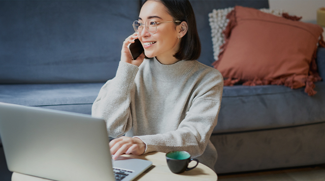 Eine junge Frau mit Brille und schwarzen schulterlangen Haaren im grauen Pulli sitzt vor der Couch an einem Couchtisch aus Holz. Auf diesem steht eine dunkelgrüne Kaffeetasse und ein aufgeklappter Laptop. An diesem arbeitet die Frau mit der rechten Hand. In der linken Hand hält sie zeitgleich ein Handy.