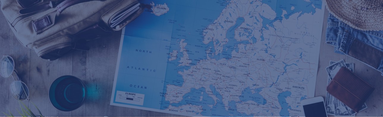 Das Bild ist blau getönt. Eine Karte von Europa liegt auf einem Holztisch neben einem Rucksack, einer Pflanze, einem Buch, einem Handy, einem Geldbeutel, einem Sonnenhut und weiteren Gegenständen.