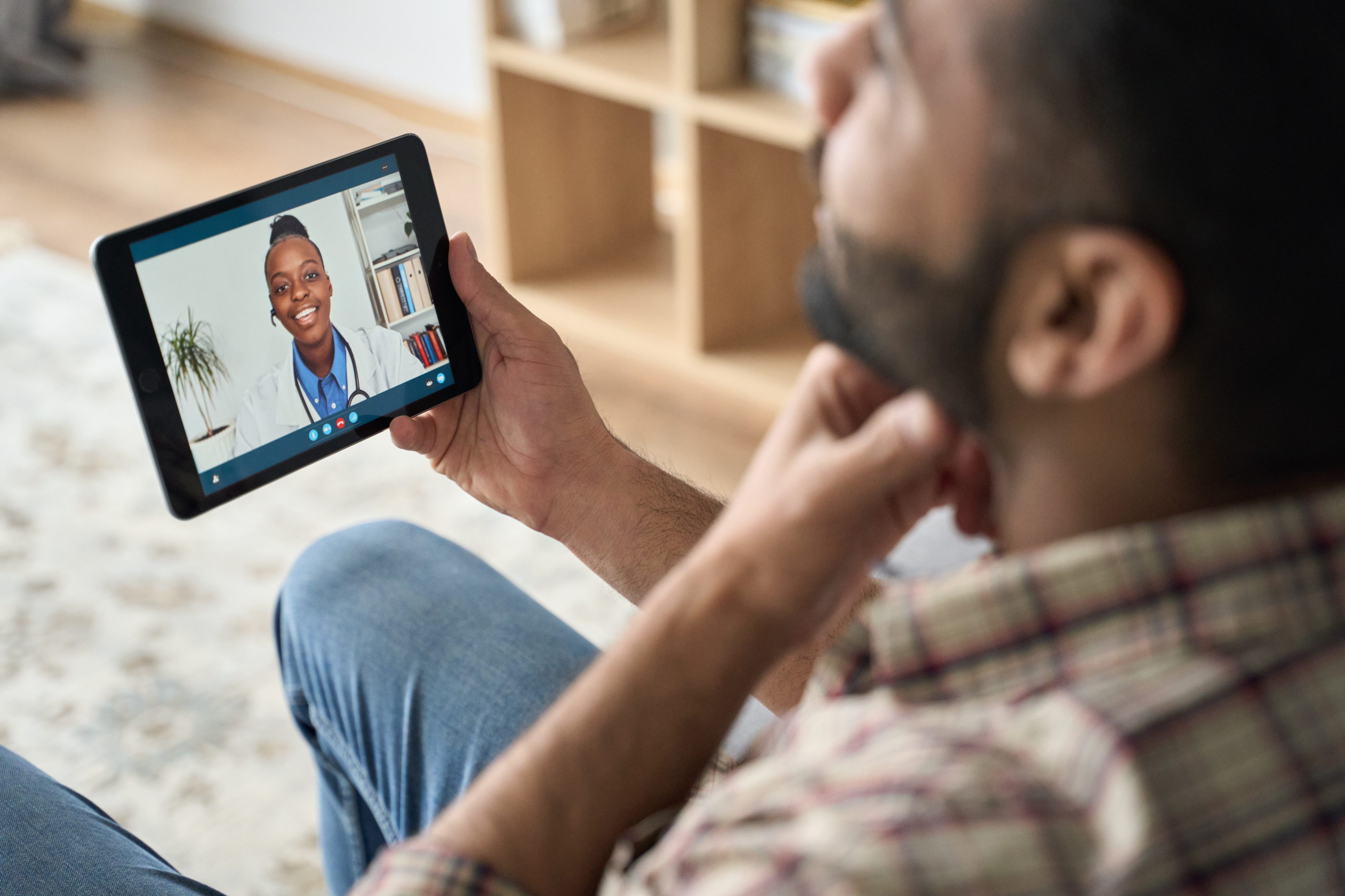 Ein Mann ist in einem Videotelefonat am Tablet mit einer Ärztin. Er sitzt im Wohnzimmer und die Ärztin lächelt.