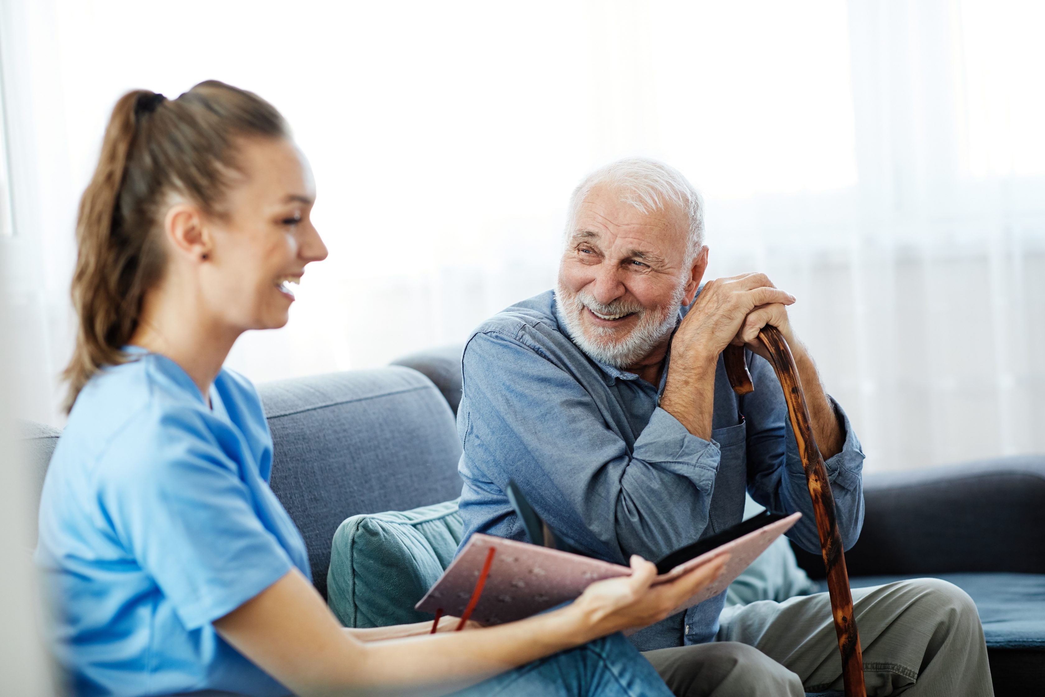 Ein alter Mann mit weißen Haaren und einem Gehstock sitzt auf einem Sofa mit einer Frau, die eine offene Mappe in der Hand hält. Beide Personen lachen.