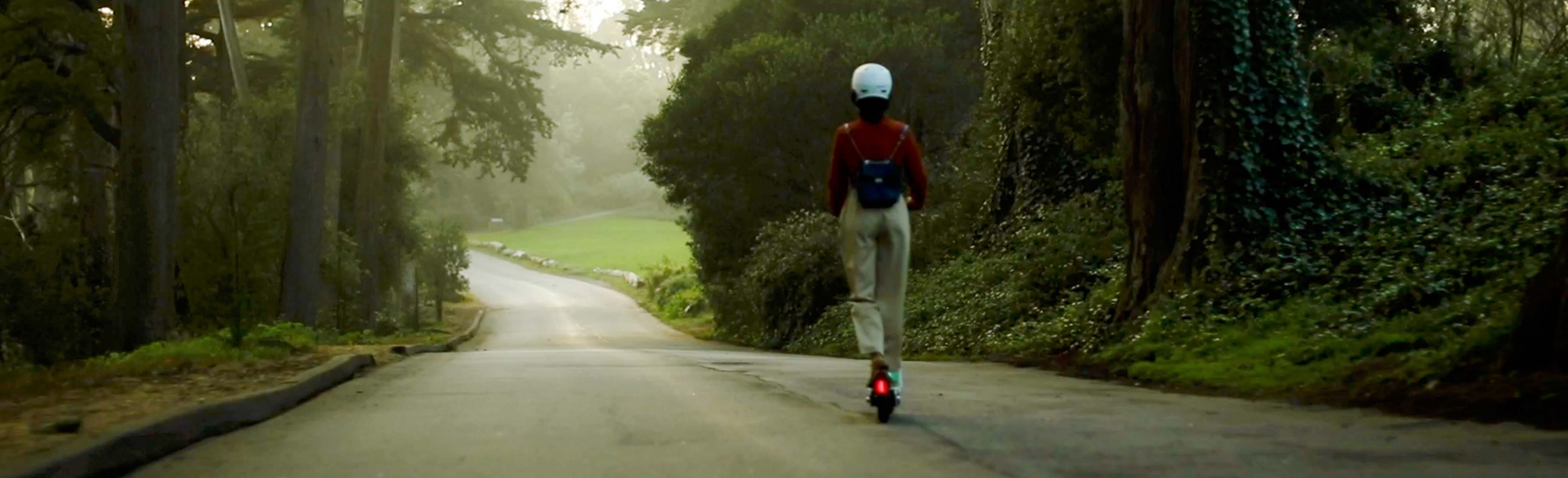 Das Bild ist blau getönt und zeigt im Hintergrund eine Person mit Helm auf einem Roller, die eine Straße im Wald entlangfährt. 