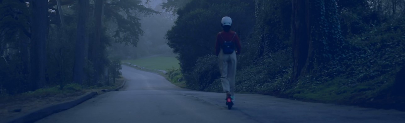 Allianz Partners Deutschland - Mobilität: Eine Frau fährt auf einem Elektroroller eine Straße durch den Wald entlang.