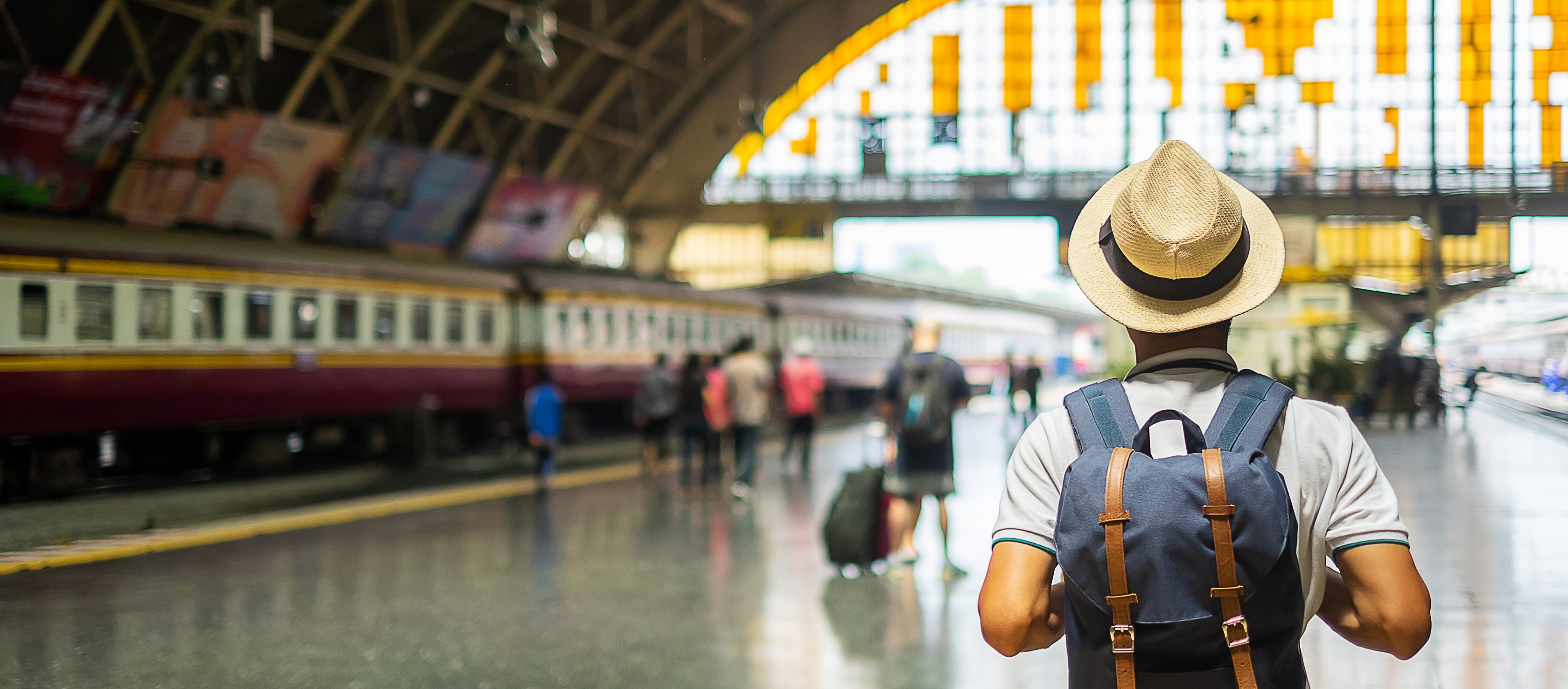 Ein Mann mit Rucksack und Sonnenhut läuft am Bahnhof. Im Hintergrund sind Gleise und Menschen zu sehen.