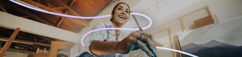 Eine Frau sitzt auf einem Sofa und schaut lachend auf ihr Smartphone. Im Hintergrund ist eine Zimmerpflanze auf einem Tisch.