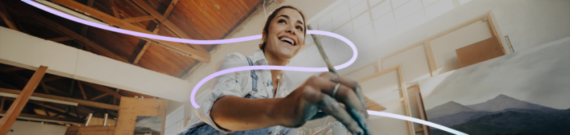 Eine Frau mit farbverschmierten Händen hält einen Pinsel in der Hand und lacht. Im Hintergrund sind Gemälde zu erkennen, sowie ein fliederfarbener, geschwungener Strich, welcher sich durch das Bild zieht. 