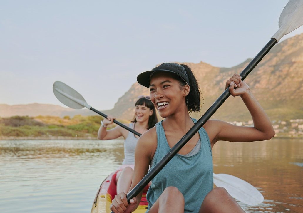Zwei Frauen sitzen im Kajak auf einem See und halten lachend die Paddel in den Händen. Im Hintergrund ist eine Berglandschaft und eine Stadt am Ufer.