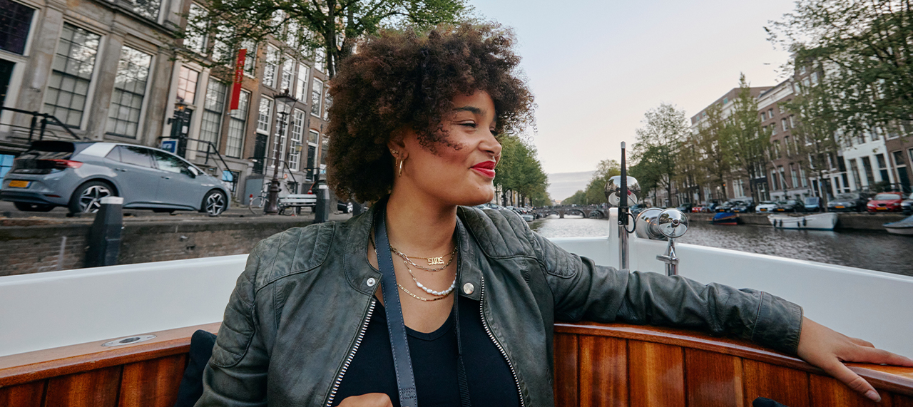 Eine Frau sitzt in einem Boot und schaut lächelnd zur Seite. Im Hintergrund ist der Kanal, Häuser und Autos zu sehen. Sie ist in Amsterdam.