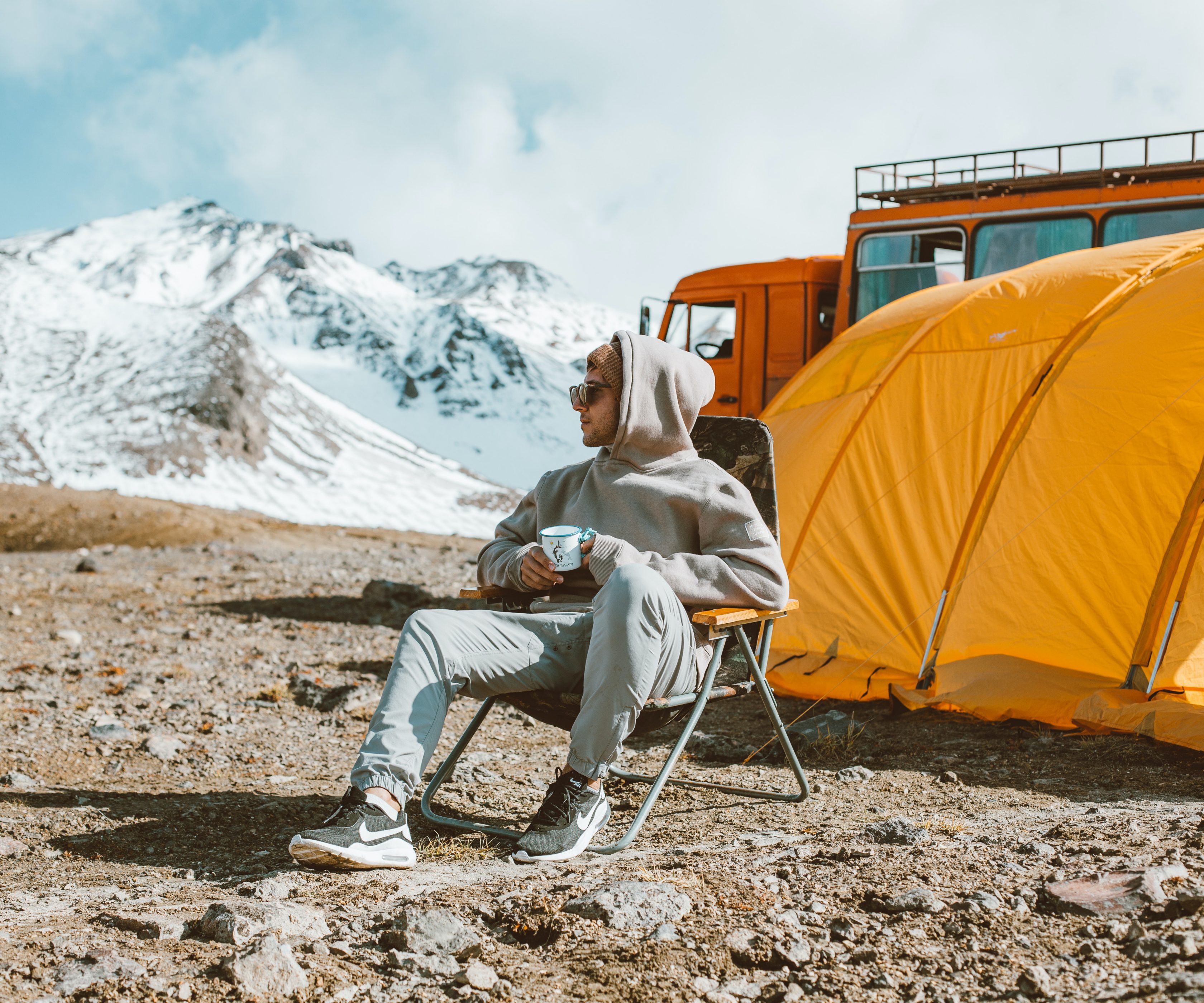 Ein Mann mit Sonnenbrille und kapuzenpulli sitzt auf einem Campingstuhl vor einem orangenen Zelt und schaut lächelnd zur Seite. Im Hintergrund sind schneebedeckte Berge und ein orangener Bus zu sehen.