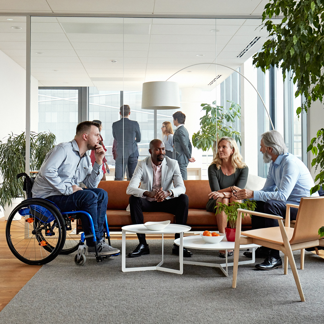 Eine Gruppe an menschen sitzen auf einem Sofa um einen Tisch herum und unterhalten sich. Im Hintergrund ist ein Büroraum mit Pflanzen und großen Fensterfronten zu sehen sowie weitere Menschen in Anzügen, die sich unterhalten.