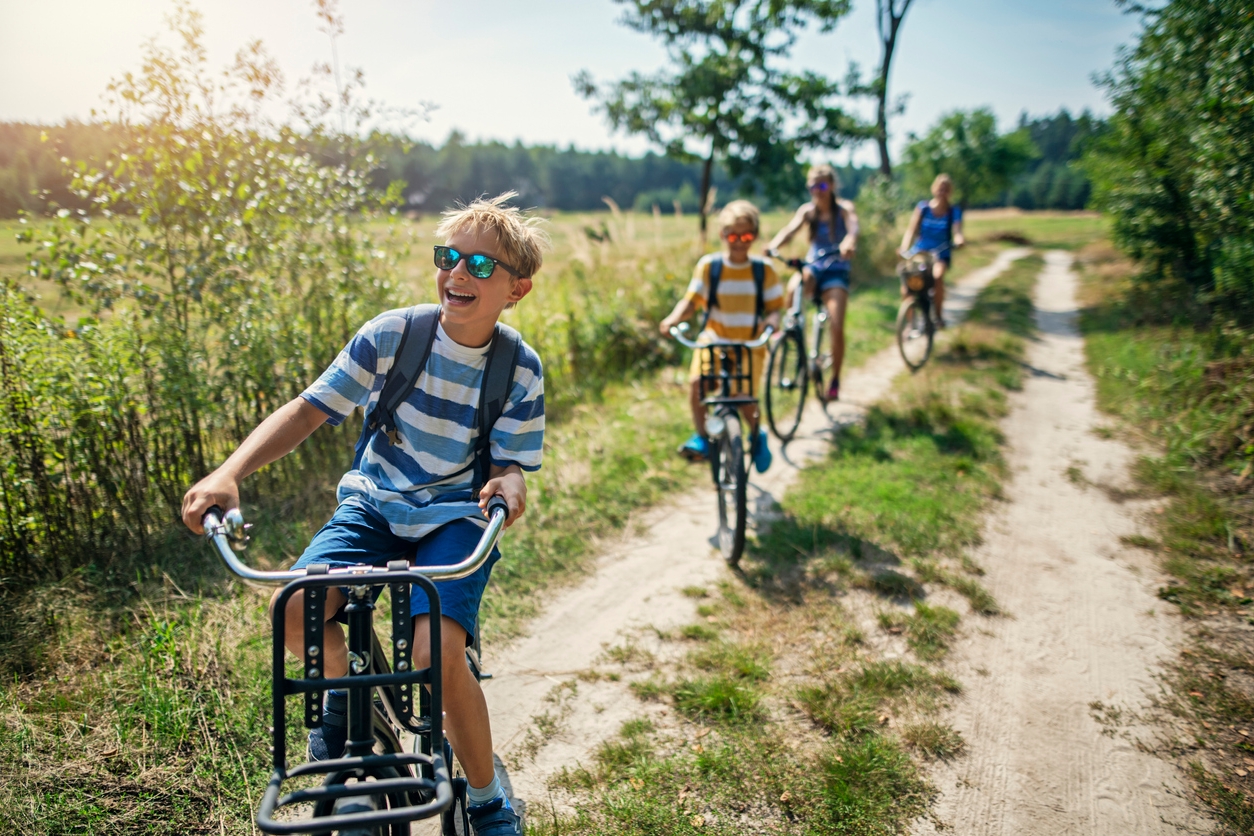 Ein Kind fährt lachend Fahrrad an einem sonnigen Tag auf einem Feldweg. Im Hintergrund ist noch ein Kind und zwei Erwachsene auf dem Fahrrad zu erkennen, die lachen.