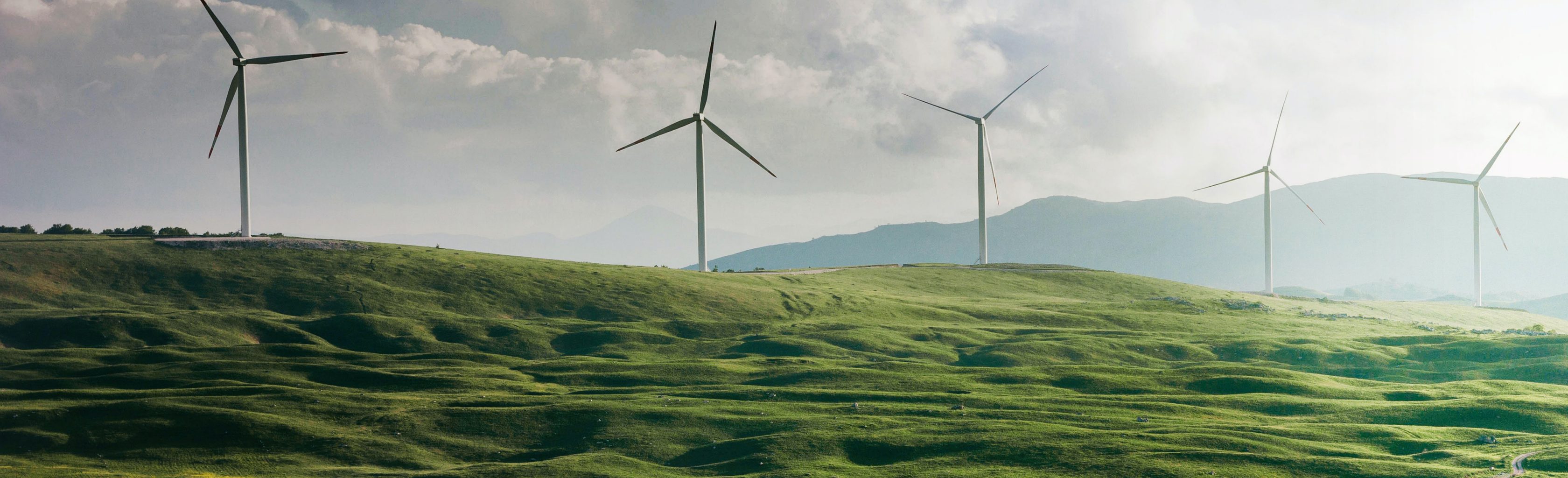 Fünf Windräder stehen auf einer grünen, hügeligen Landschaft. Im Hintergrund sind mehr Berge und Hügel sowie ein Wolkenhimmel zu sehen.
