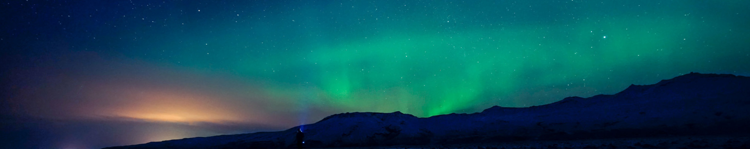 Allianz Partners Deutschland - Über uns: Eine Person mit Taschenlampe steht vor einer dunklen Berglandschaft, während das grüne Polarlicht den Nachthimmel erhellt.