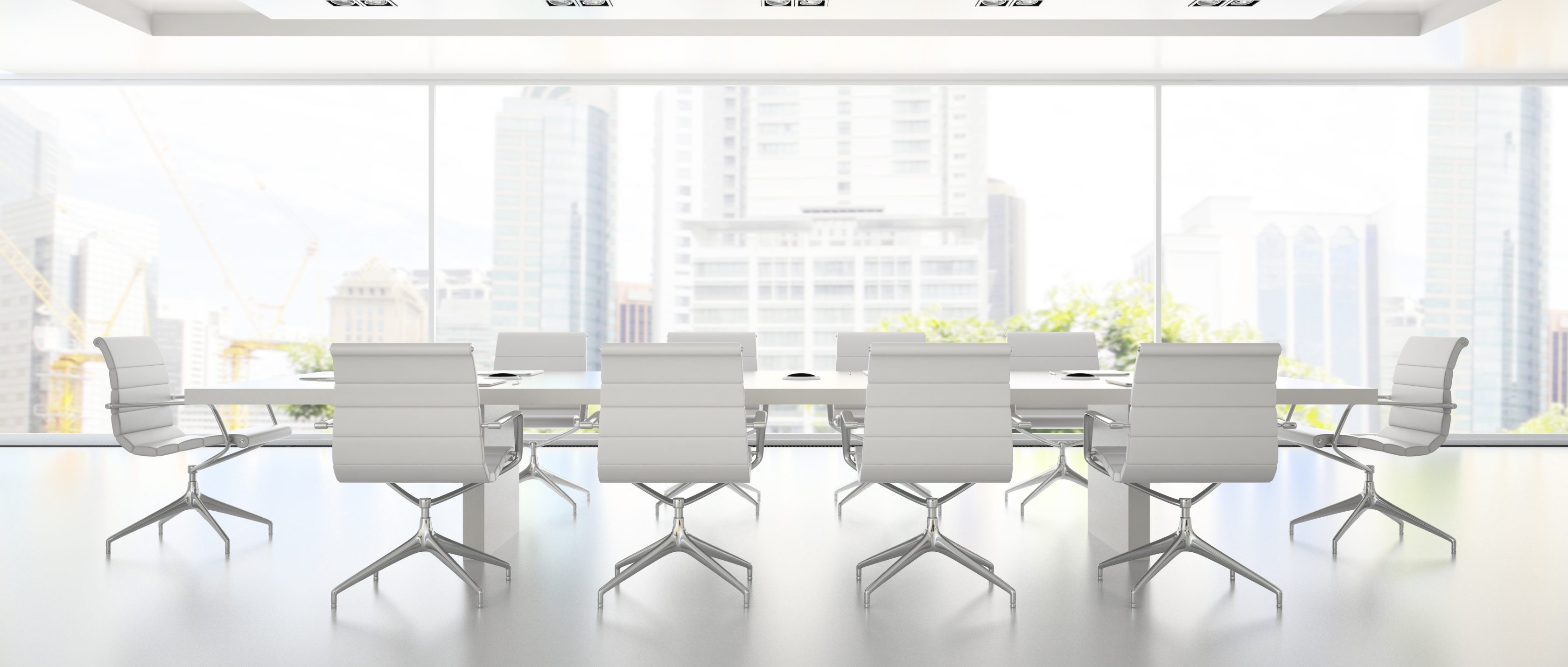 Das Bild zeigt es sehr helles Besprechungszimmer. Im Hintergrund ist eine Fensterfront mit einer Stadtsilouhette zu sehen. Im Raum selbst steht ein langer Tisch mit zehn Tischen, davon je vier an den langen Seiten und einer an den kurzen Seiten. Alles ist in weiß gehalten und daher strahlend hell.