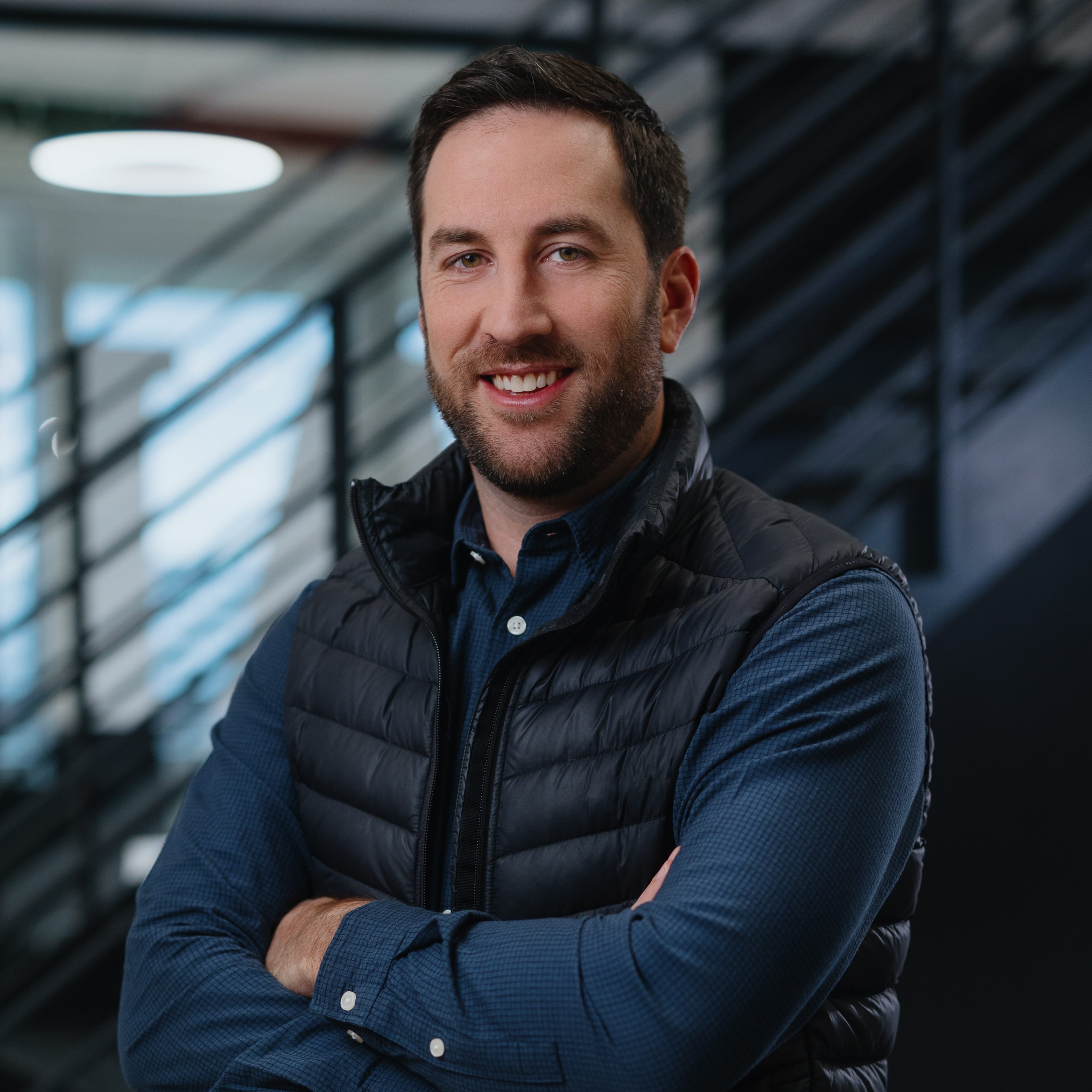 Porträt Phil Hoffman, Chief Travel Officer bei Allianz Partners. Mann mit dunklem Haar, schwarzer Weste über dunkelblauem Hemd, lächelt in die Kamera und verschränkt die Arme vor der Brust.