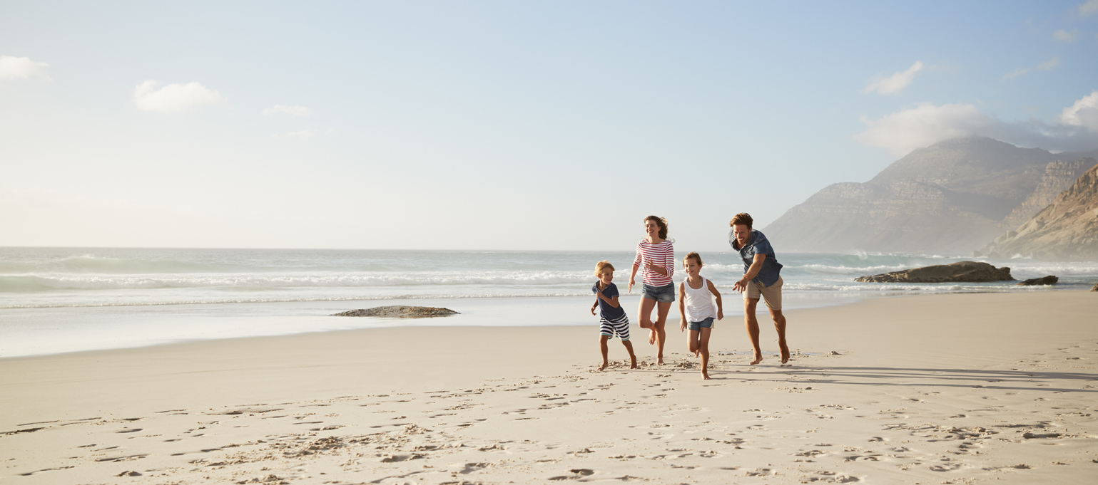 Família correndo na praia. Uma mulher e um homem estão atrás e um menino e uma menina estão na frente. Todos estão com roupas para o verão.