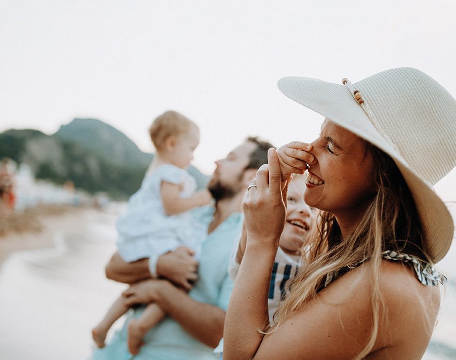 Eine Familie mit zwei Kindern sind am Strand. Die Frau hält ein Kind auf dem Arm, das der Frau an Nase greift. Frau und Kind lachen.
