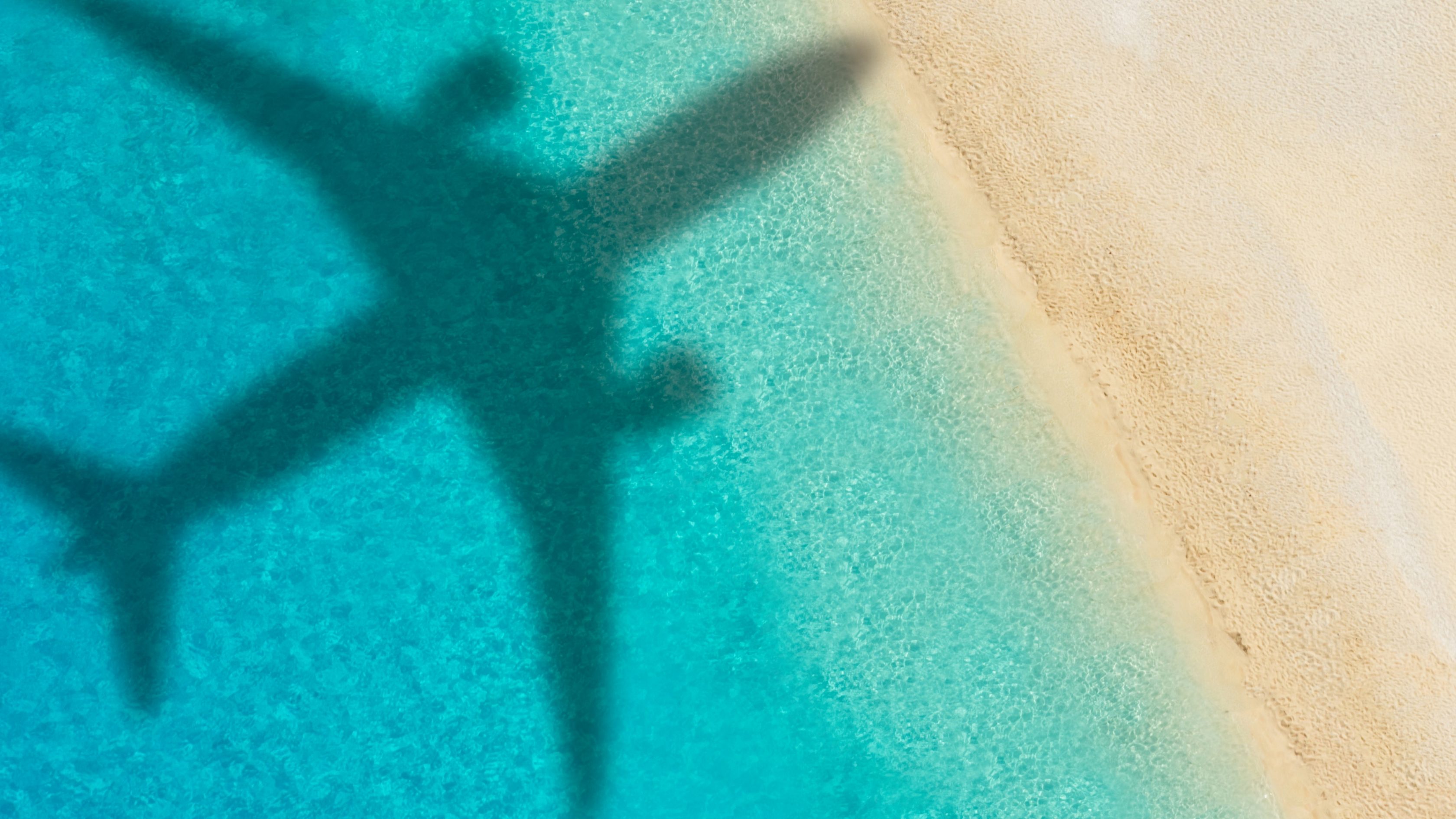 Der Schatten eines Flugzeuges ist auf dem türkisen Wasser an einem Sandstrand mit grünen Palmen zu sehen.