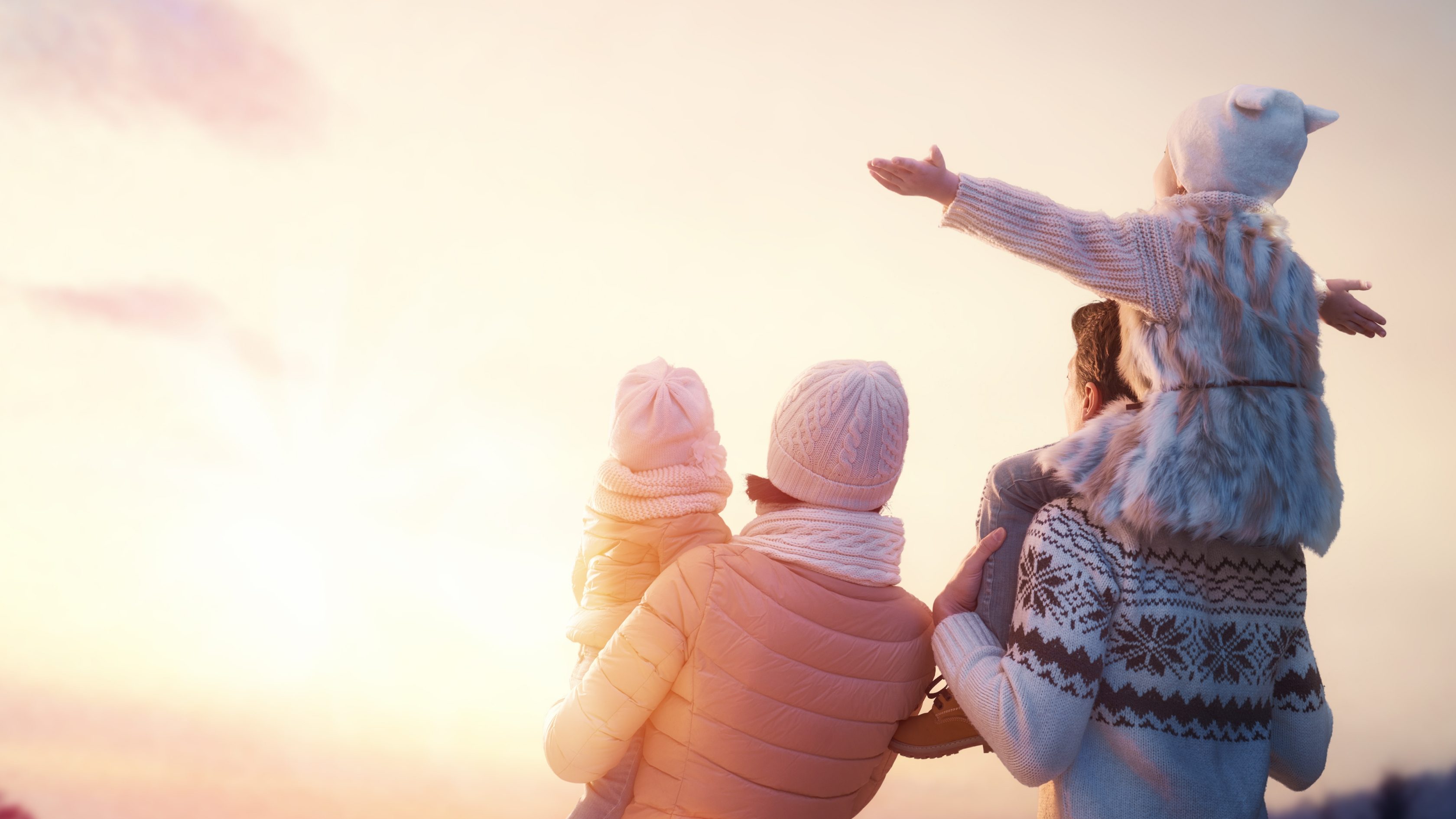Eine Familie mit zwei Kindern schauen dem Sonnenuntergang am Himmel an. Ein Kind sitzt auf den Schultern des Vaters, das andere trägt die Mutter im Arm.