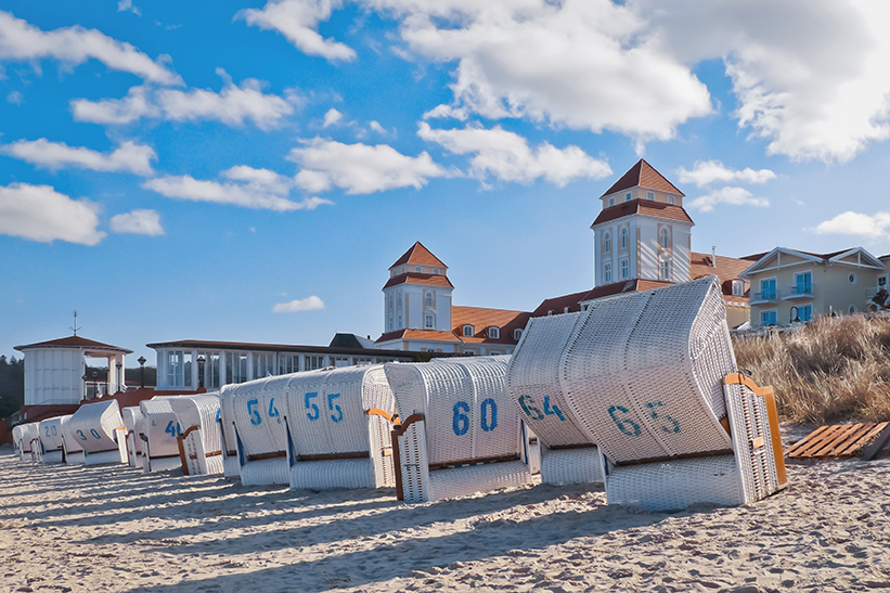 Weiße Strandkörbe zum Sitzen stehen an einem Sandstrand. Im Hintergrund sind Gebäude mit roten Dächern und ein Wolkenhimmel zu sehen.