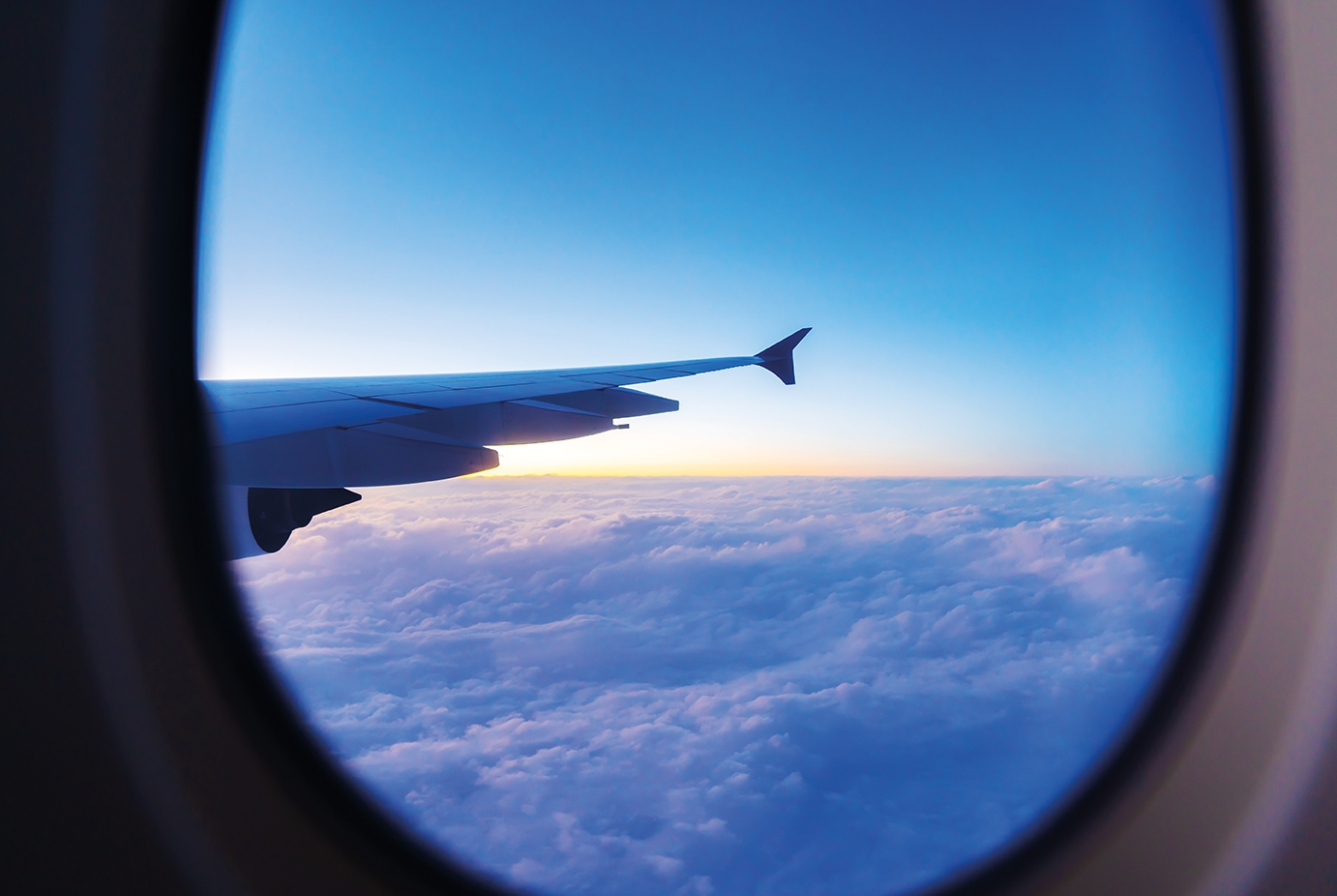 Aus einem ovalen Flugzeugfenster ist ein Flugzeugflügel über einer Wolkendecke am gelb-blauen Horizont.
