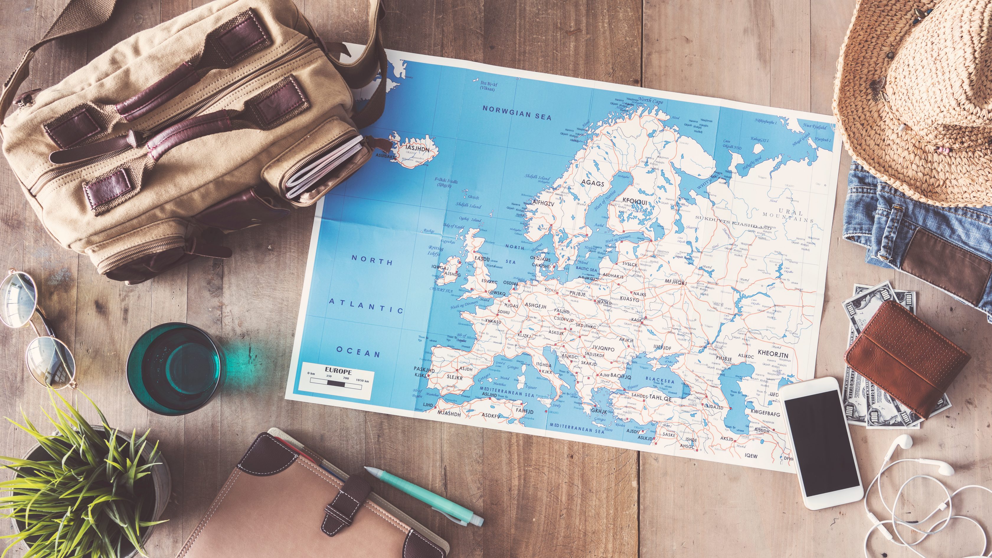 Eine Landkarte von Europa liegt auf einem Holztisch. Daneben sind eine Pflanze, Bücher, ein Smartphone, Kopfhörer, ein Hut und ein Rucksack zu sehen.