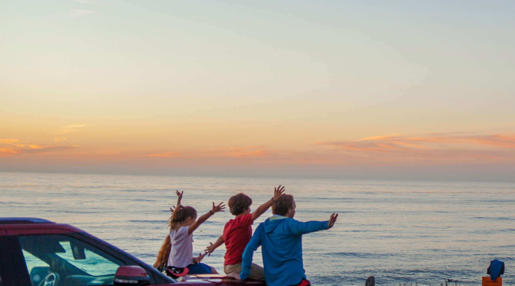 Eine Familie sitzt an einem Auto und blickt auf das Meer vor ihnen bei Sonnenuntergang. Die Kinder heben vor Freude die Arme hoch.