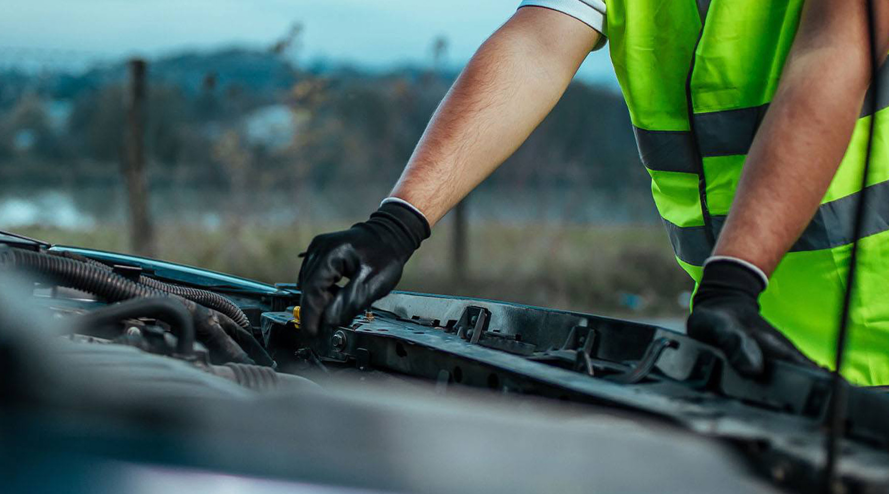 Eine Person mit Warnweste und Handschuhen arbeitet an einer geöffneten Motorhaube eines Autos.